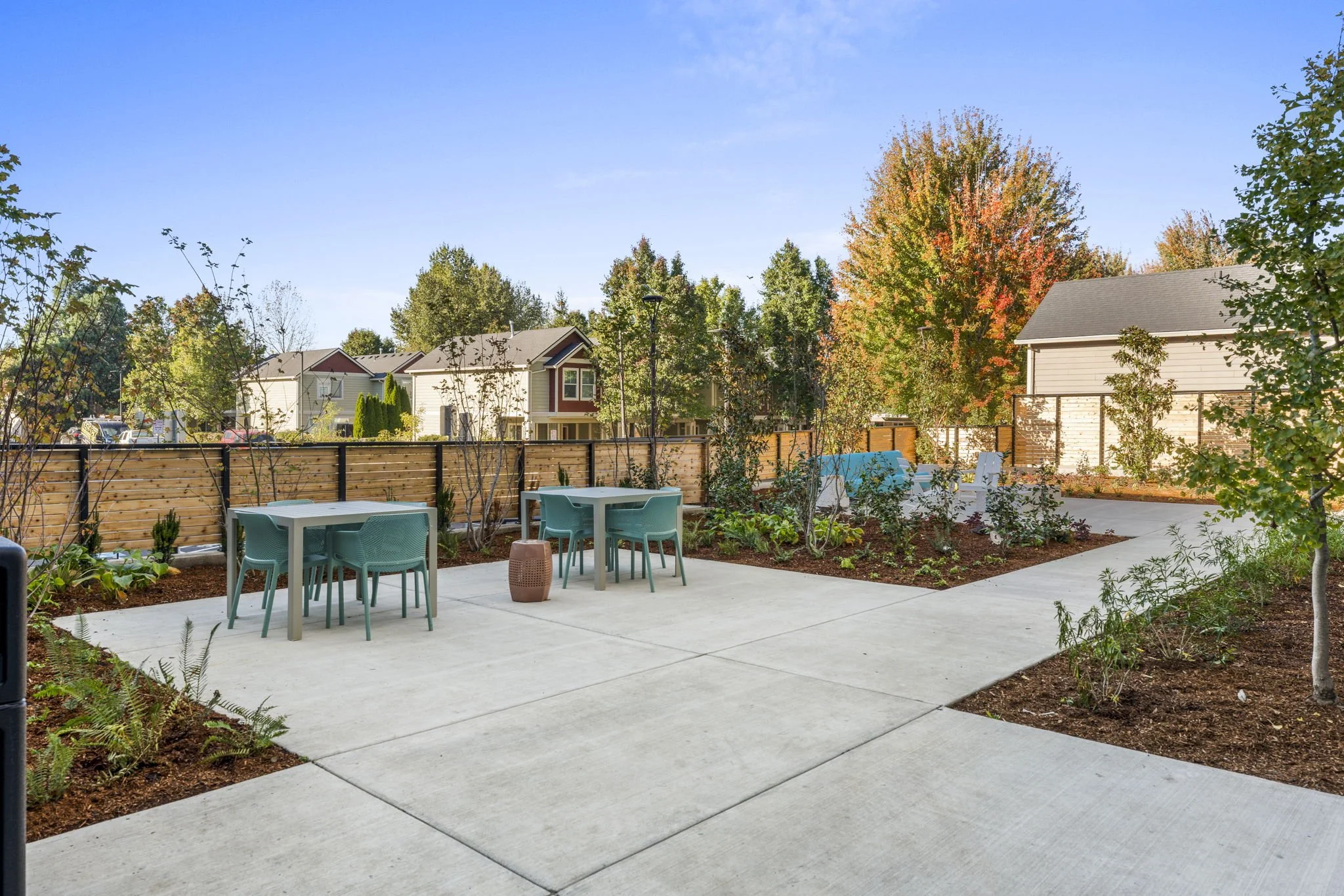 Backyard patio with outdoor tables, chairs, and surrounding plants, with houses and trees in the background under a clear blue sky.