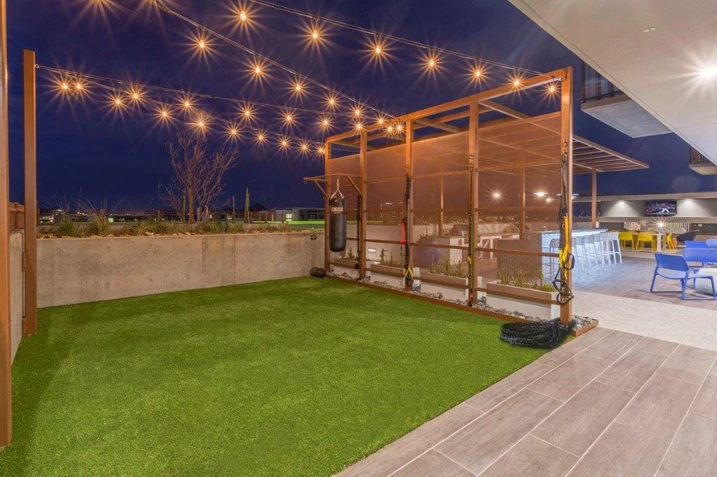 Outdoor space at night with string lights, a grassy mat, and a wooden fitness structure with boxing bags and resistance bands. There are chairs and tables in the background under a covered area.