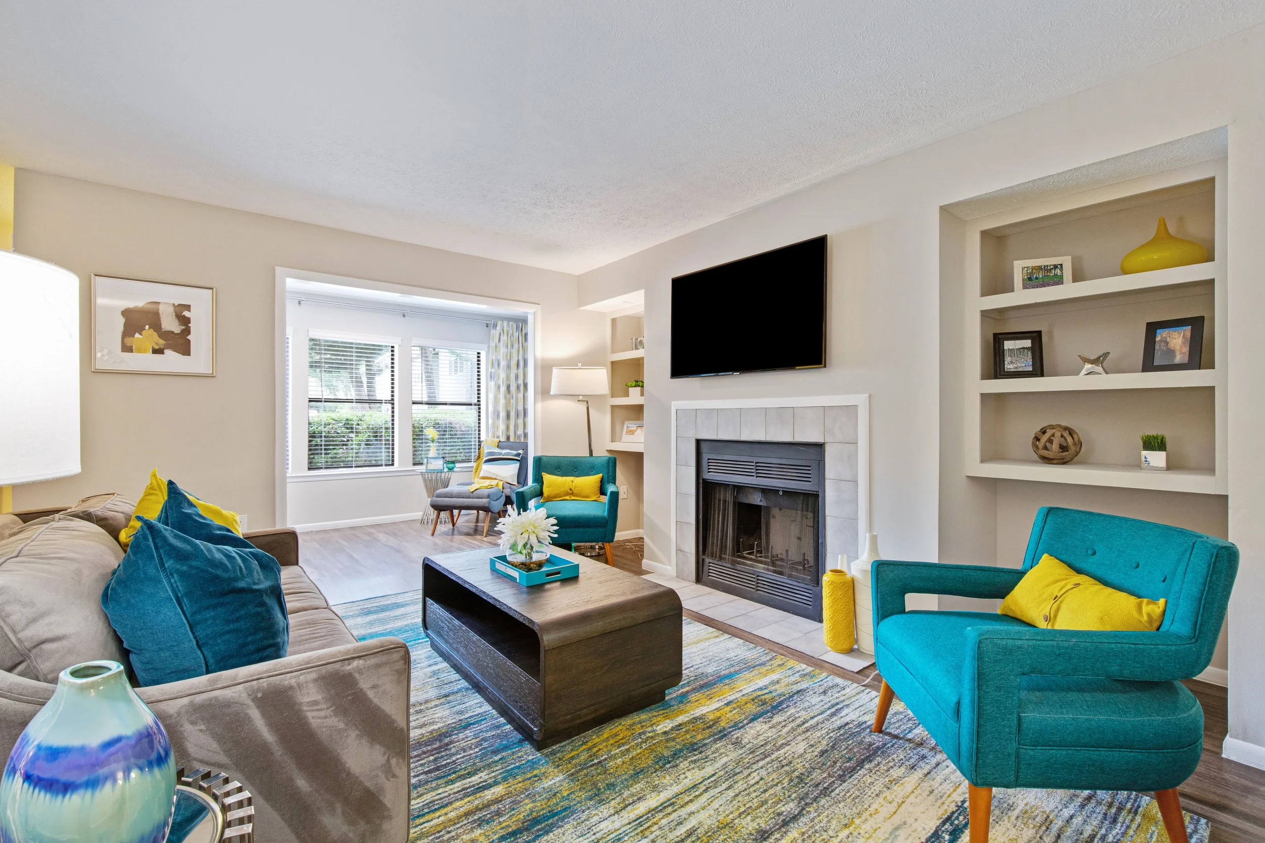 Living room with blue and yellow accent chairs, a beige sofa with blue and yellow pillows, a wooden coffee table, a fireplace with a mounted TV, and built-in shelves with decorative items, colorful rug, large window with curtains.