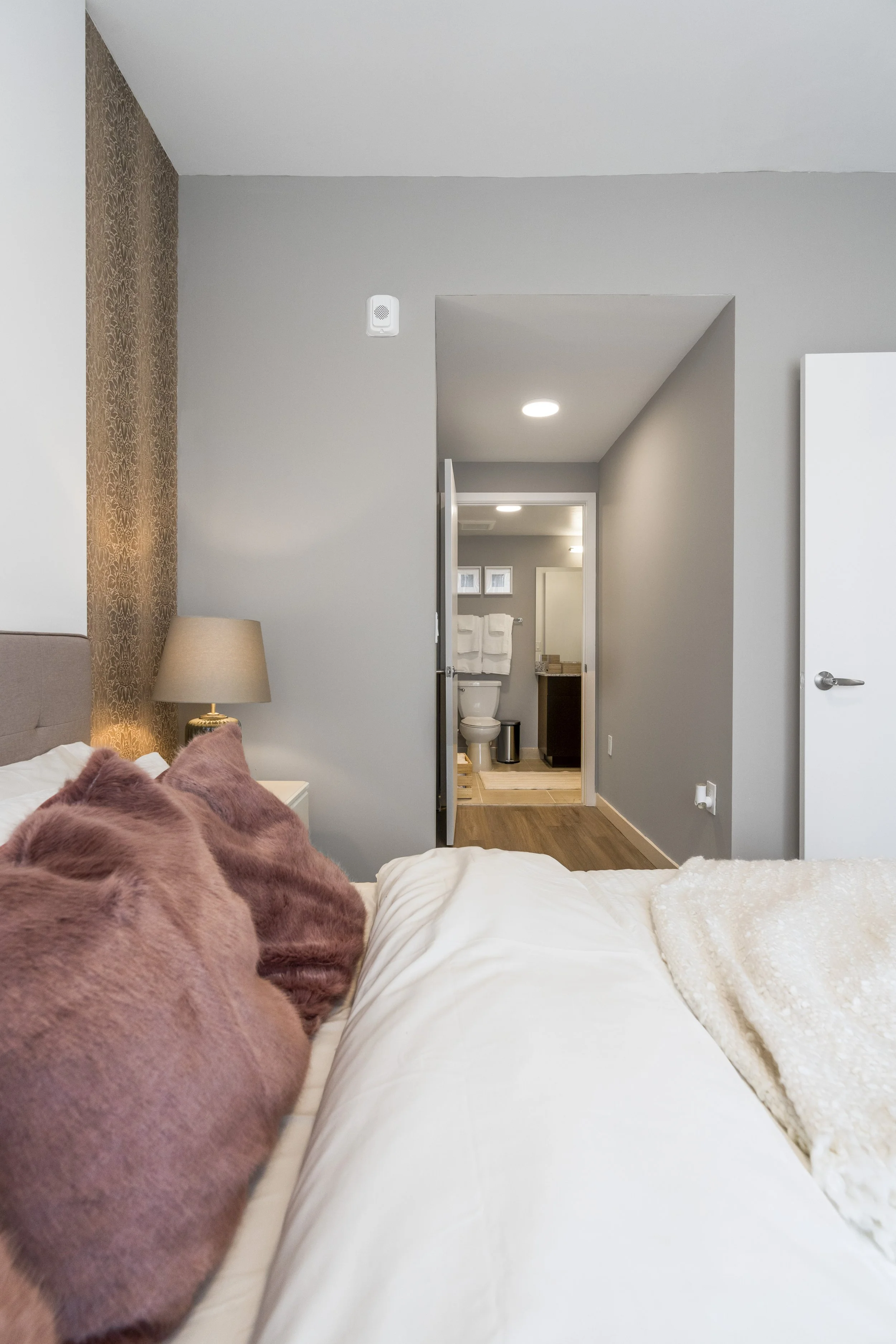 A bedroom with a bed, pink pillows, a nightstand with a lamp, and a view of a bathroom through an open door.