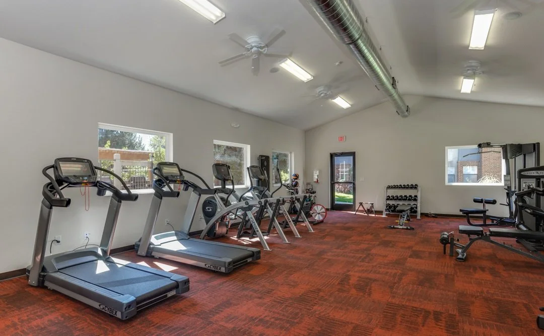 Empty fitness room with treadmills, elliptical machines, dumbbells, weight rack, and exercise equipment, lit by fluorescent lights and windows, with a door at the back.