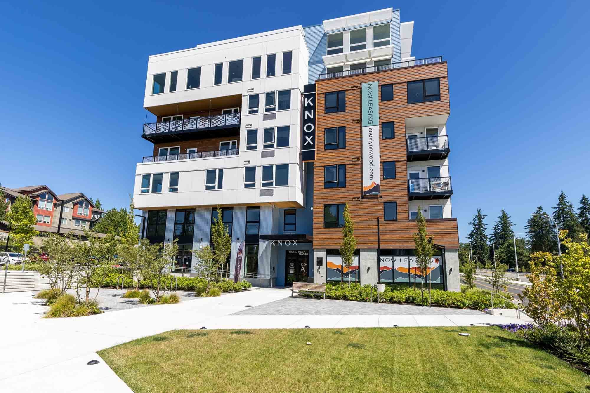 Modern multi-story apartment building with a white and wooden facade, balconies, and large windows, surrounded by landscaped greenery and a clear blue sky.