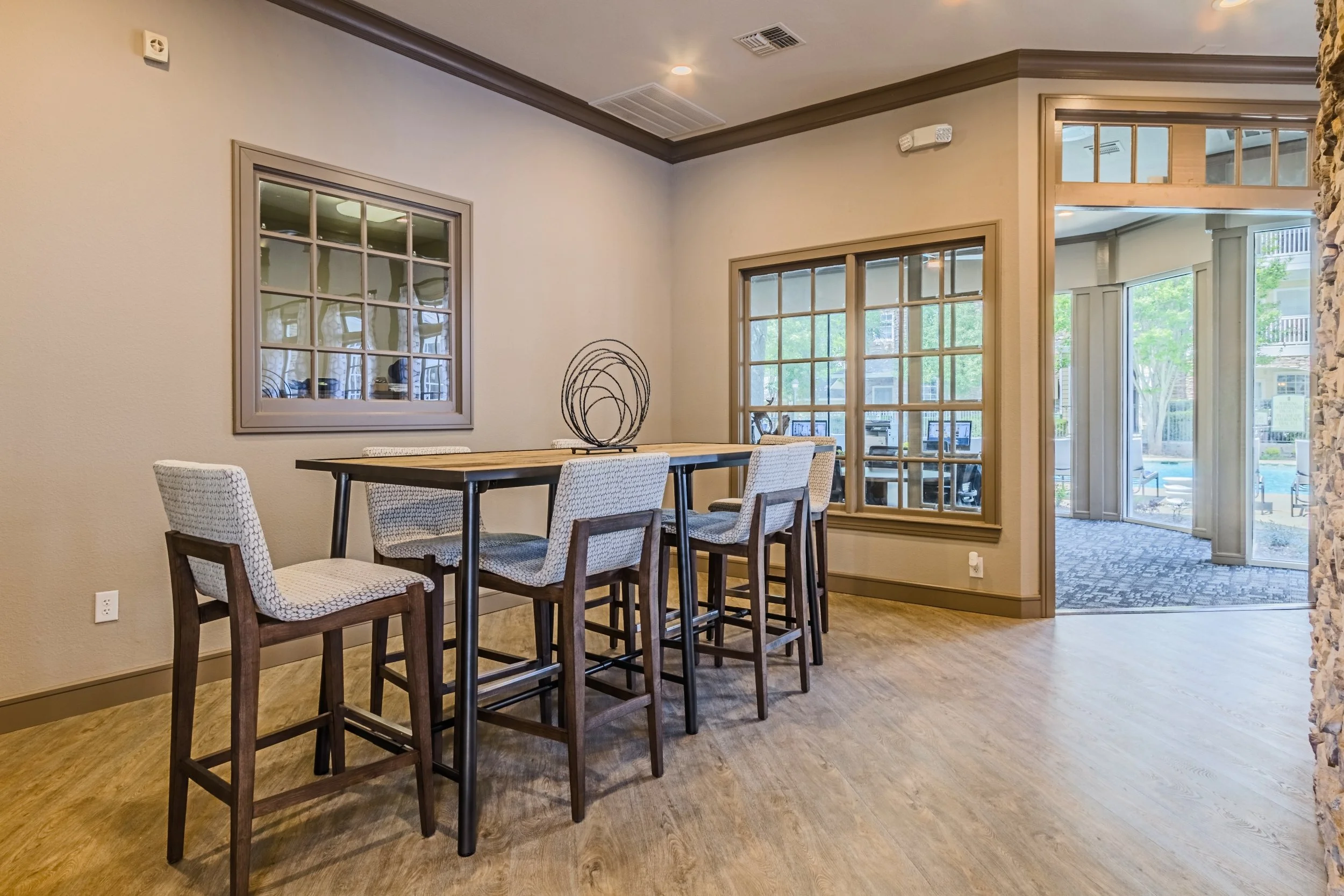 An indoor room with a high wooden table and four chairs, two with cushioned seats, near two large windows. Outside the windows, there is a view of a street with trees, buildings, and outdoor seating, as well as a sliding glass door leading to an outd