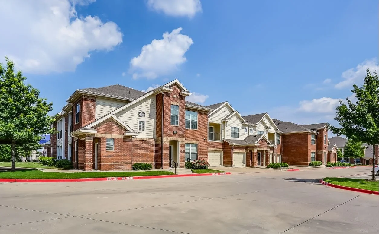 A residential apartment complex with brick and siding exteriors, multi-story buildings, and attached garages, under a partly cloudy sky.