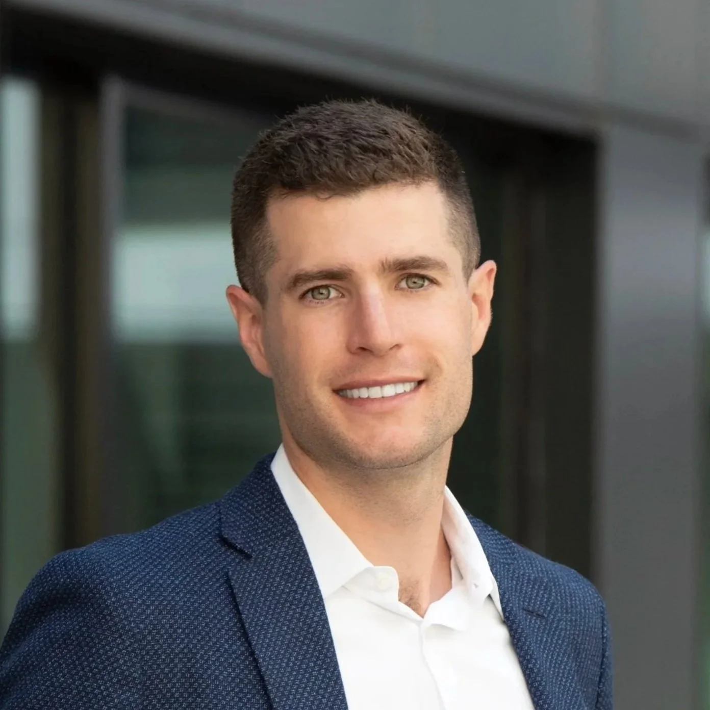 Headshot of a young man with short brown hair, blue eyes, and a smile, wearing a white shirt and a dark blue blazer, outdoors with blurred glass building in background.