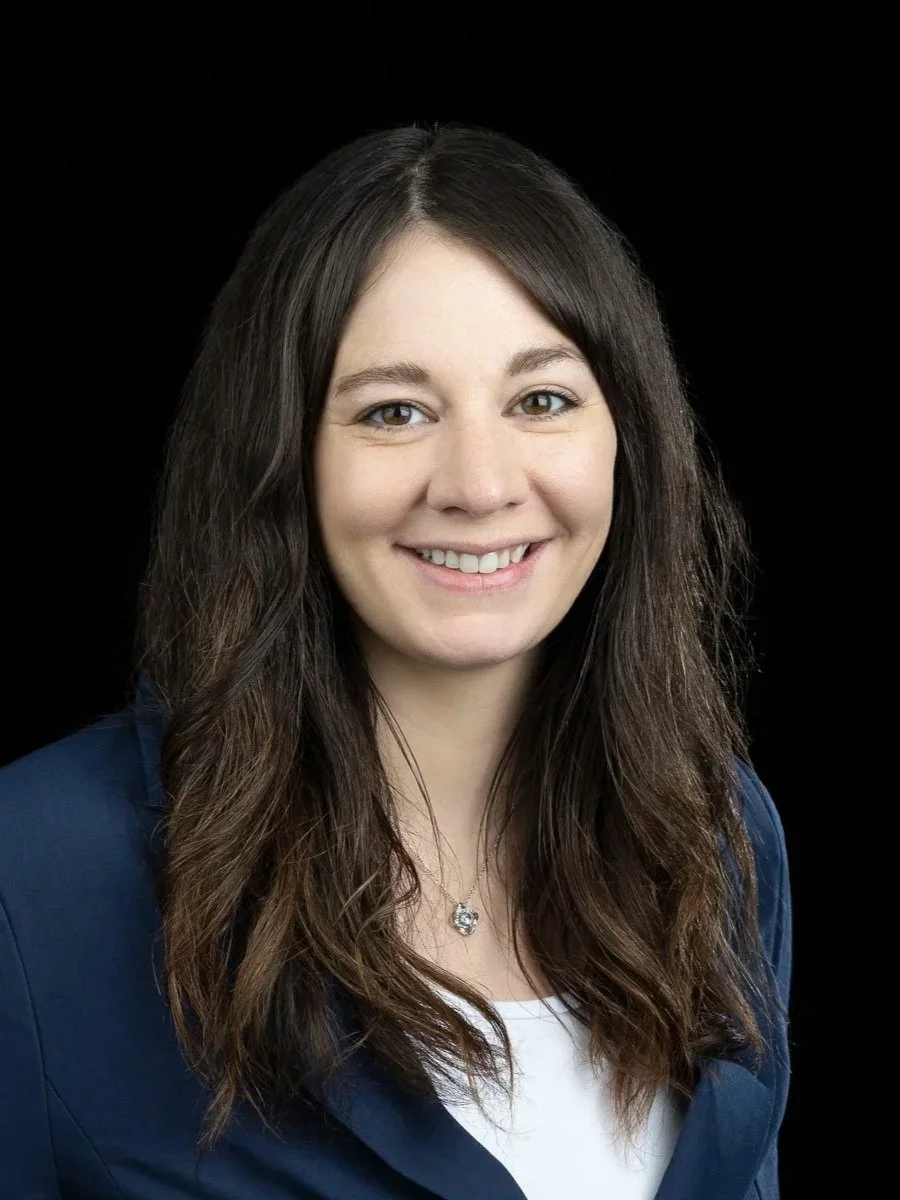 A woman with long brown hair, smiling, wearing a navy blazer and a necklace, against a black background.
