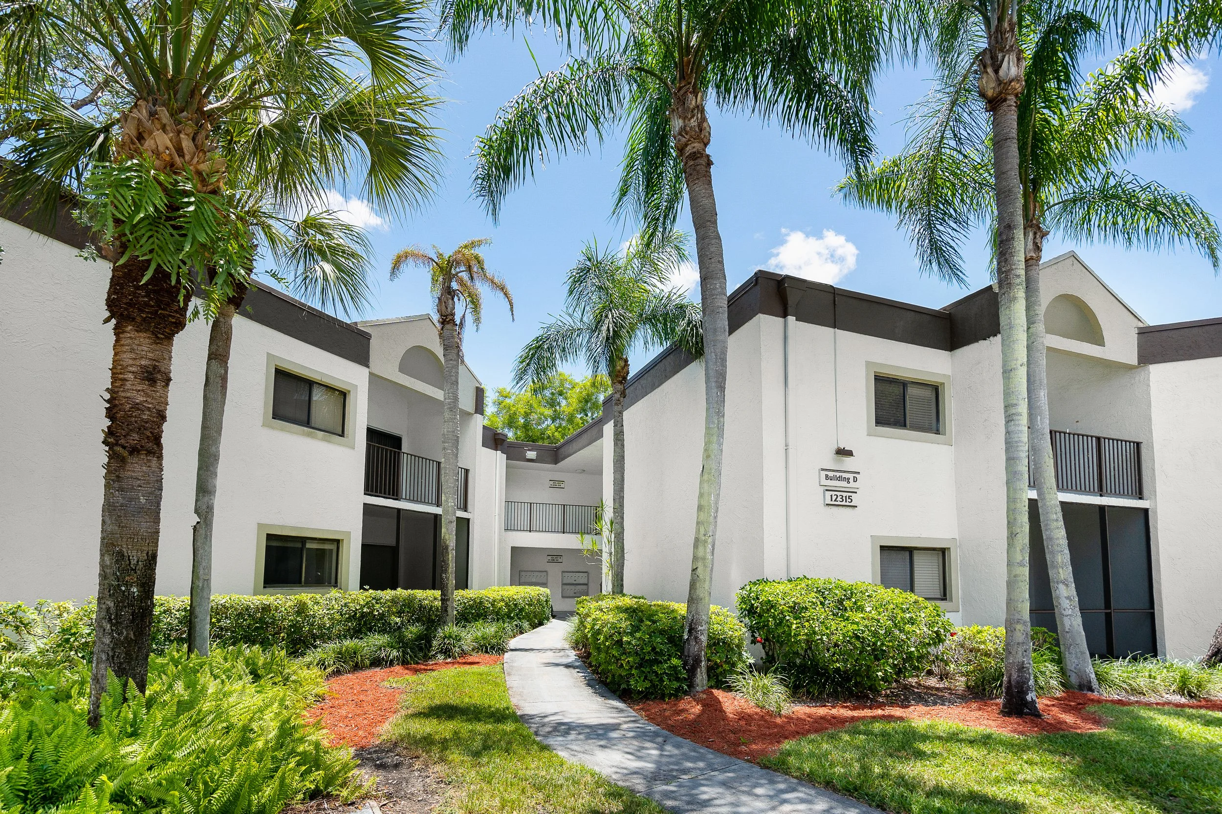 Exterior view of a modern apartment complex with white stucco walls, palm trees, a paved walkway, and lush green landscaping under a blue sky.