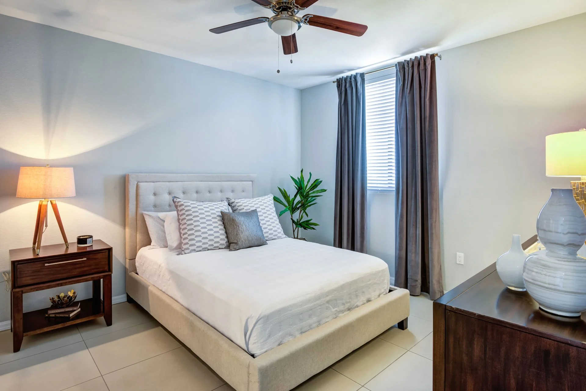 A bedroom with a beige tufted bed, gray pillows, and a plant next to a window with gray curtains. There is a wooden nightstand with a lamp and a small clock, and a dark wooden dresser with white vases. The ceiling fan has brown blades and a white lig