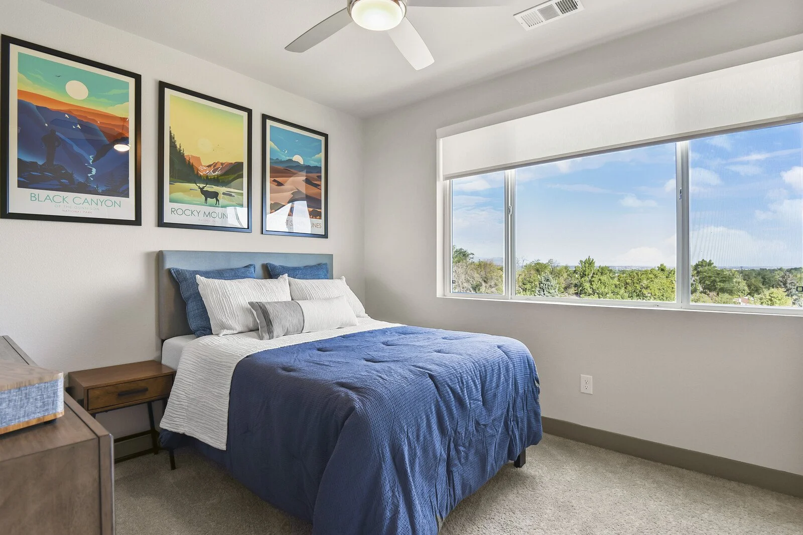Bedroom with blue bedspread, pillows, window with a view of trees and sky, and three framed landscape posters on the wall.
