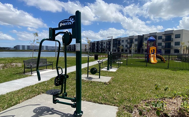 Outdoor gym equipment near a pond with a playground in the background, apartment buildings, benches, and a paved walkway under a partly cloudy sky.