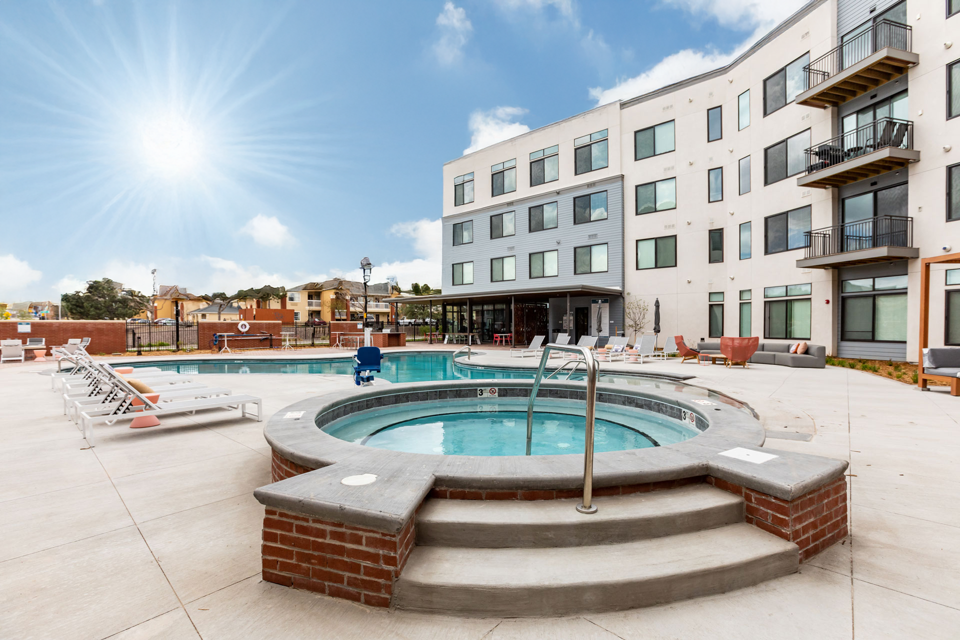 Outdoor pool area with hot tub, lounge chairs, and apartment building in daytime with sunny sky.