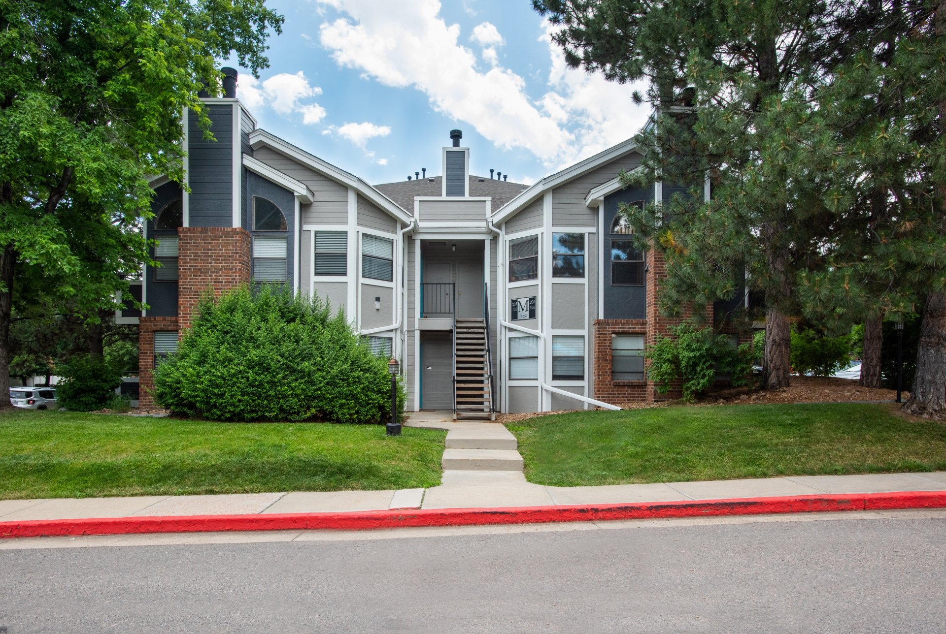 Exterior view of a multi-story apartment building with brick and siding walls, surrounded by green trees and grass, under a partly cloudy sky.