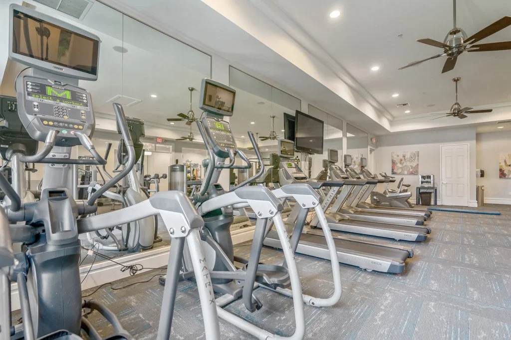 A row of exercise bikes and treadmills in a modern gym with large mirrors, ceiling fans, television screens, and paintings on the walls.