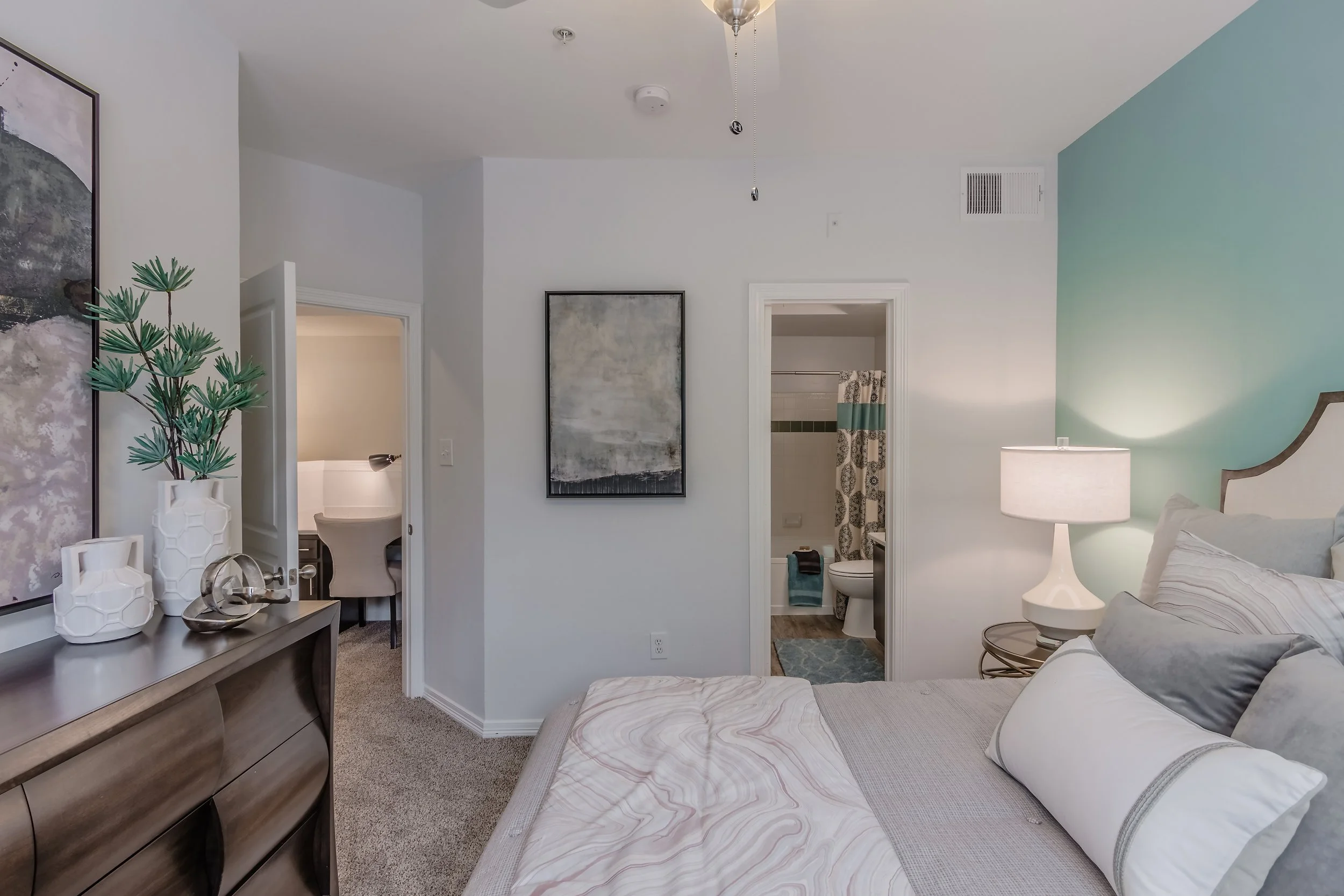 A bedroom with a bed, bedside lamp, wall art, and two bathrooms visible in the background.