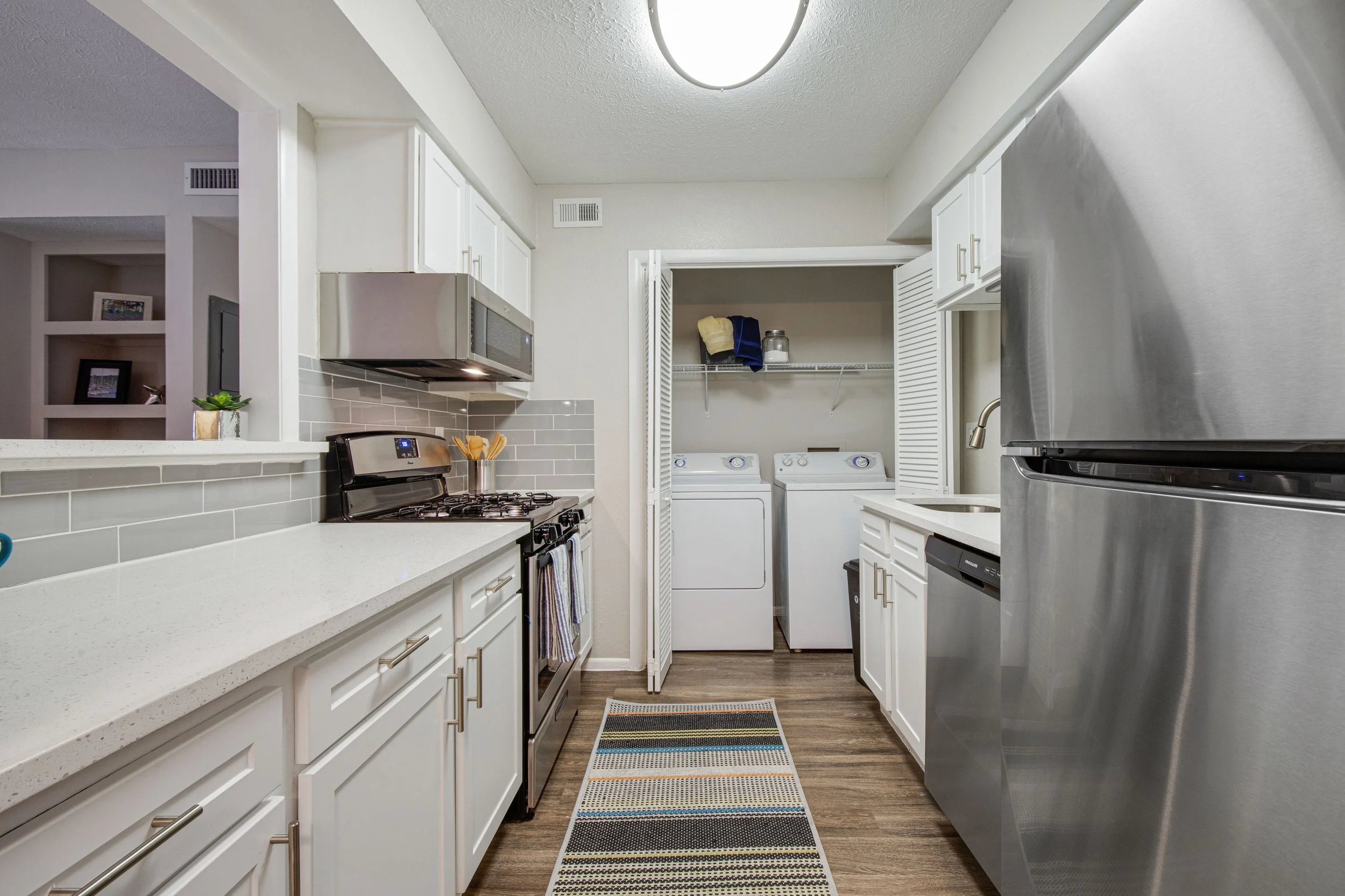 Kitchen with white cabinets, stainless steel refrigerator, microwave, stove, and a laundry area with washer and dryer. A striped rug on wood floor, opening to a living room area.