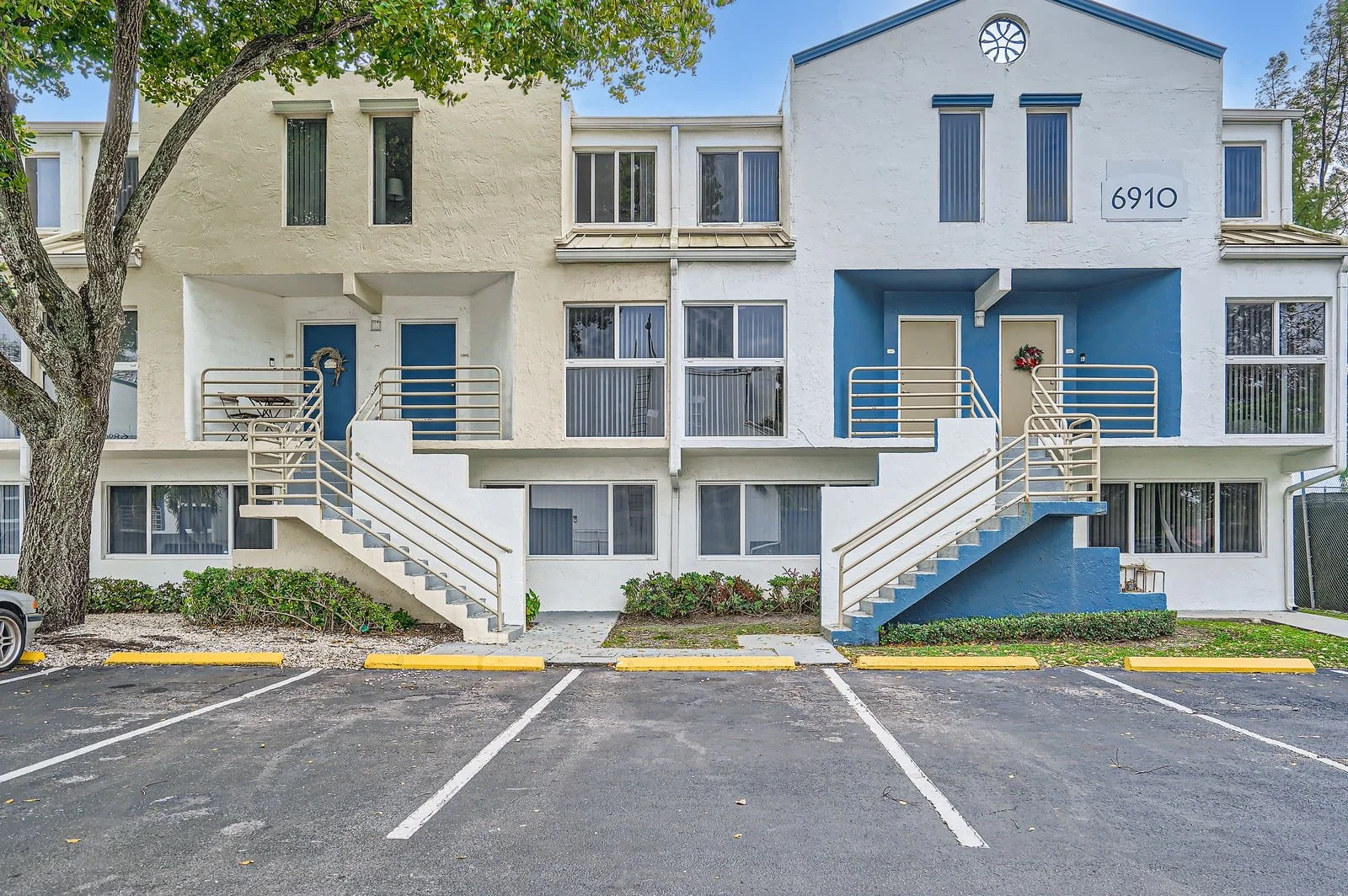 Front view of a two-story apartment building with exterior stairs leading to second-floor entrances, including a blue entrance door with a wreath and beige door. Parking spaces in the foreground, a tree on the left, and the building number 6910 on th