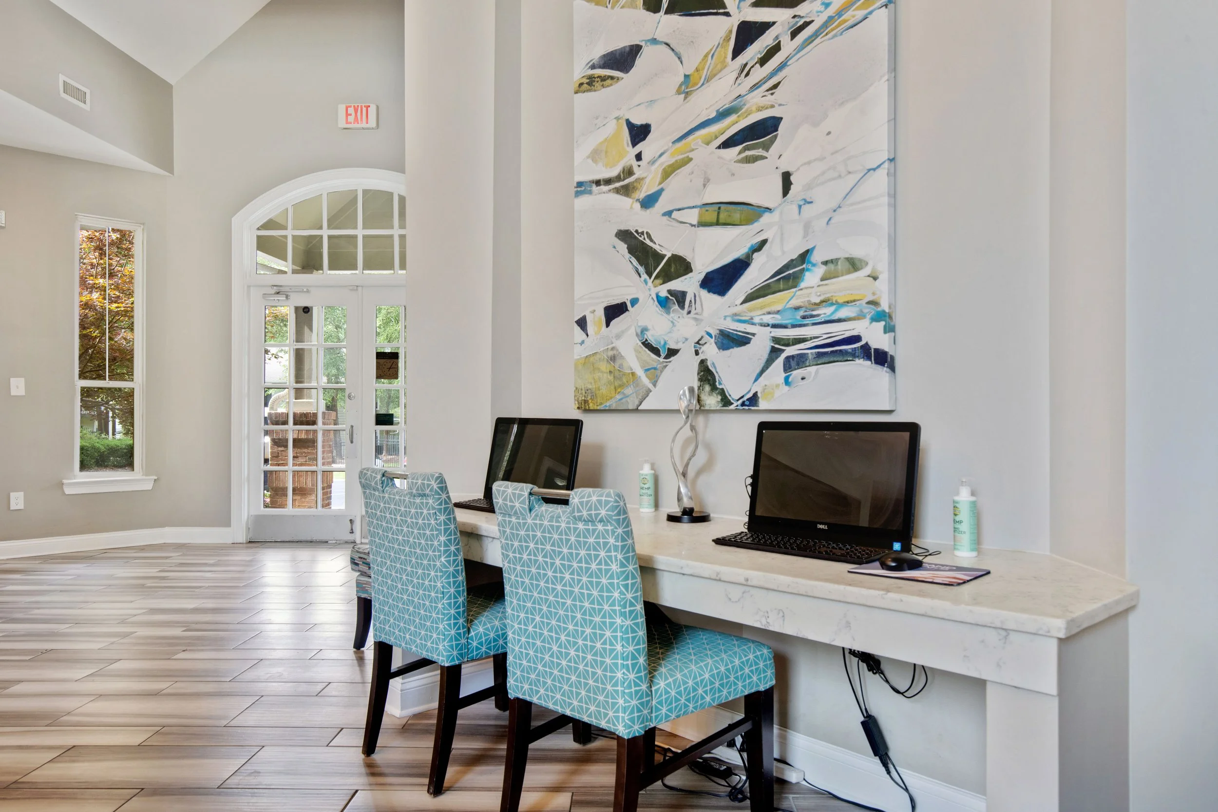 Interior view of a modern reception or work area with three yellow and green patterned chairs, and three desktop computers on a marble counter against a light wall. There is a large abstract painting hanging above the counter, and a door with a windo