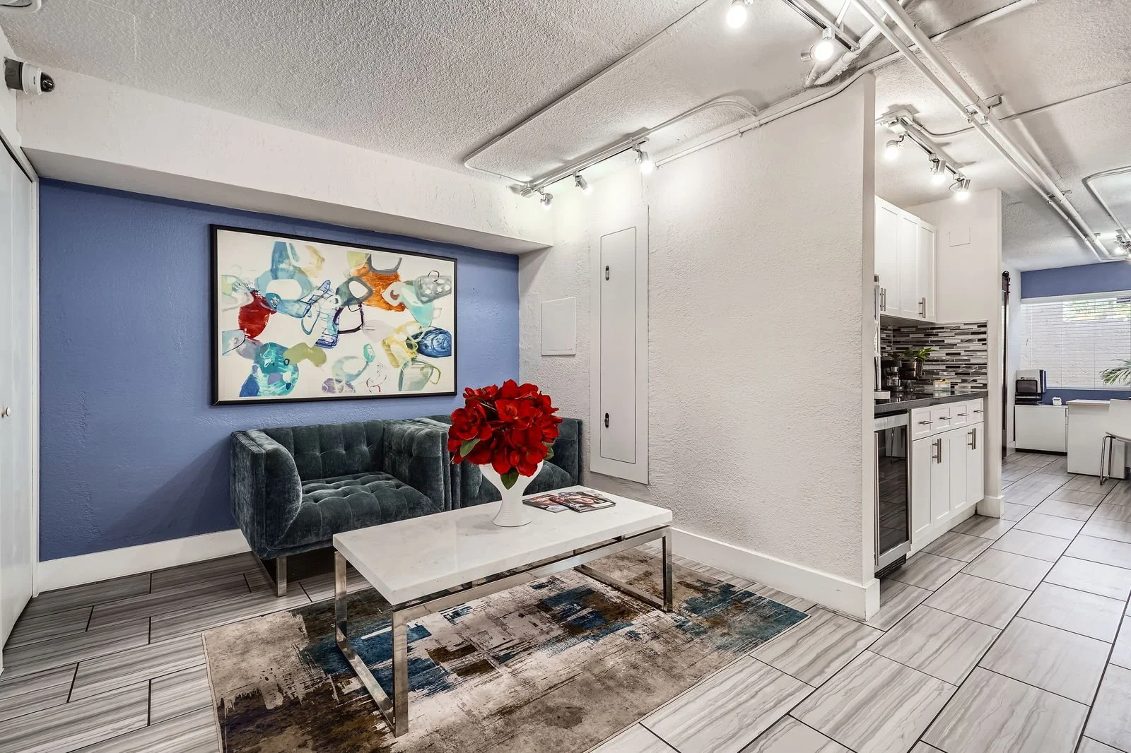 A modern living space with a dark green velvet sofa, white coffee table with a red flower arrangement, abstract artwork on a blue accent wall, and a view into the kitchen area with white cabinets and a tiled backsplash.