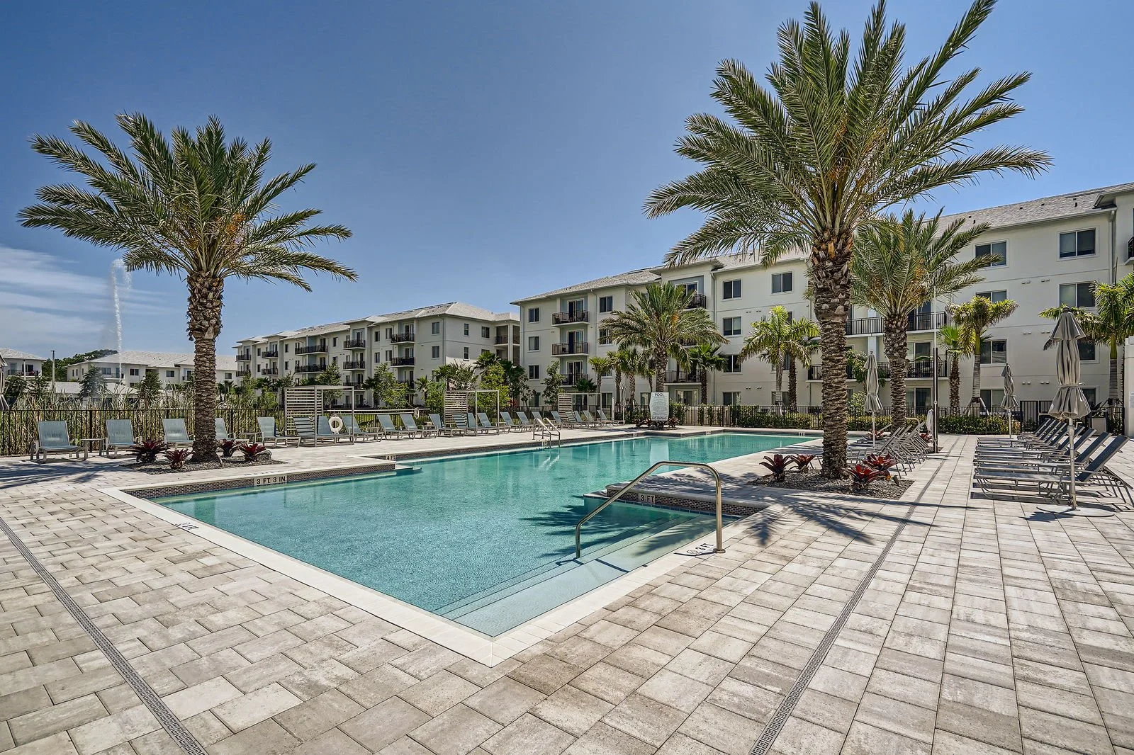 An outdoor swimming pool area with palm trees, lounge chairs, and a multi-story residential building in the background under a clear blue sky.