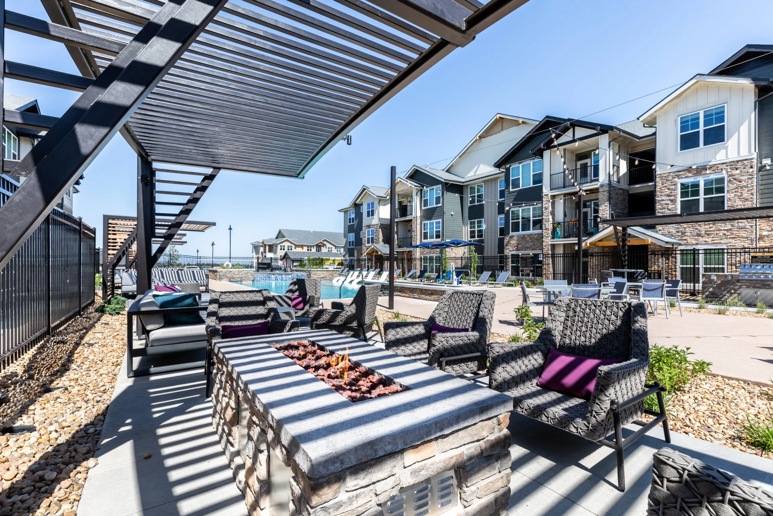 Outdoor communal area with chairs surrounding a fire pit, swimming pool, apartment buildings, and patio furniture, under a pergola in a residential complex.