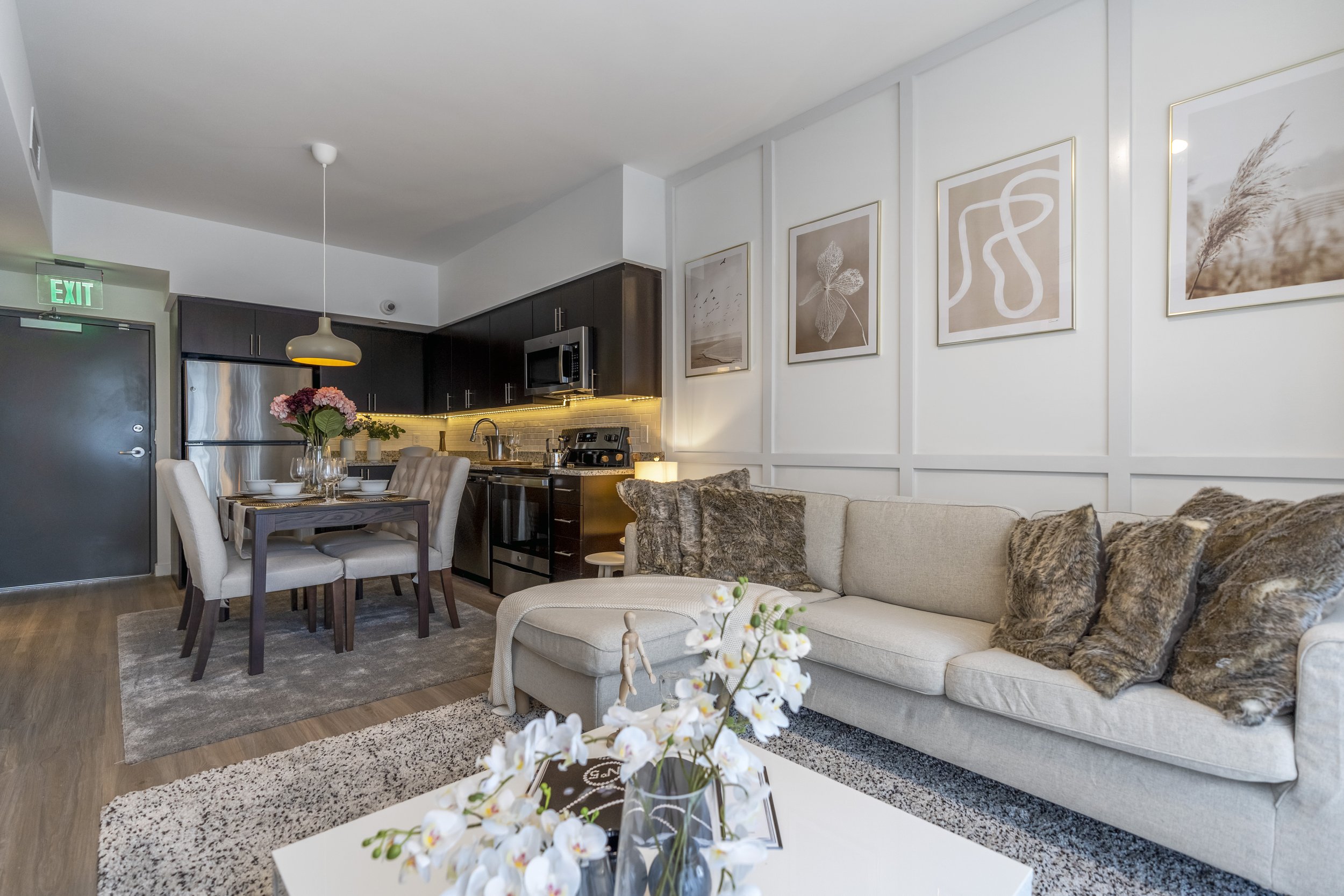 Living room and kitchen with beige sofa, patterned cushions, dining table with six chairs, flowers, and modern kitchen appliances.
