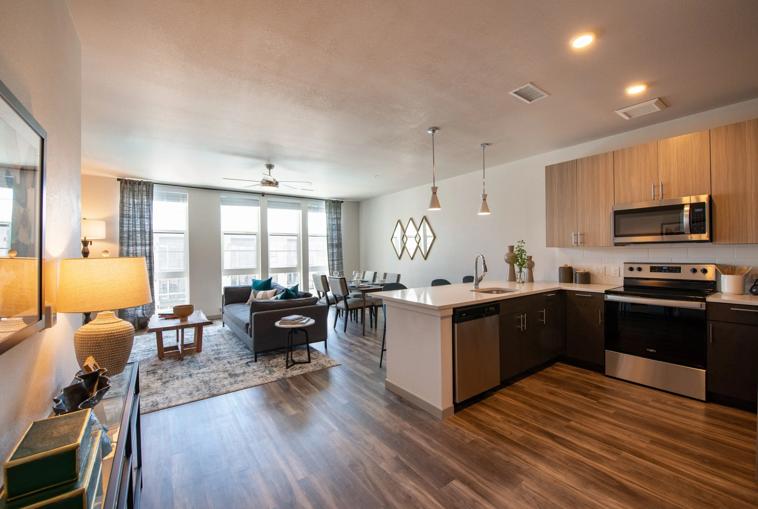 Open-concept living area with a kitchen and dining space, featuring hardwood floors, modern furniture, a sofa with cushions, large windows with striped curtains, and decorative wall art.