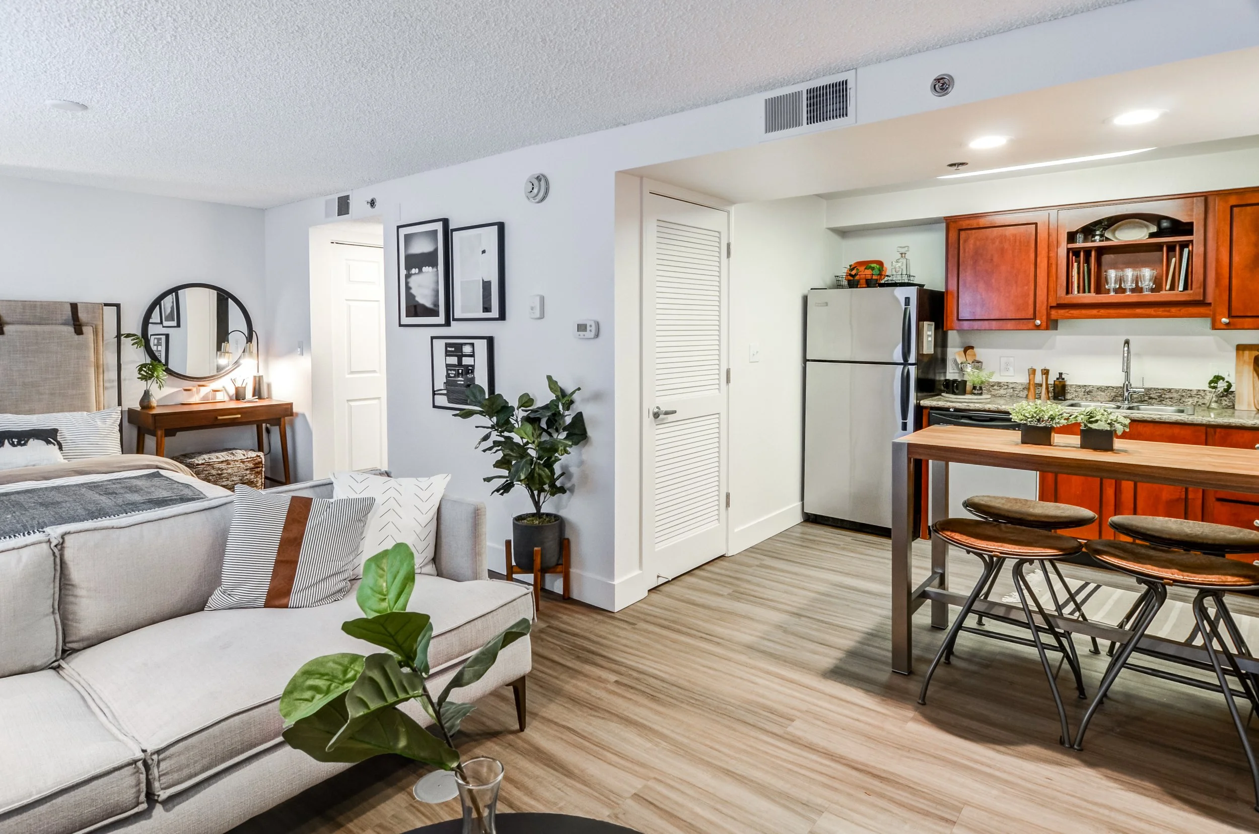 Living room and kitchen in an apartment with wooden flooring, a beige sofa, a small table with plants, a white fridge, wooden cabinets, and a breakfast bar with bar stools.