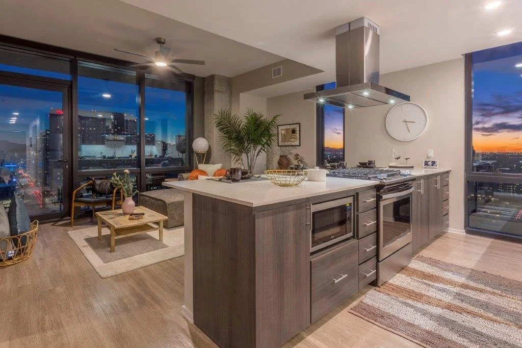 Modern open-concept apartment with kitchen and living room, city skyline views at sunset, wooden floors, contemporary furniture, large windows, and stainless steel appliances.