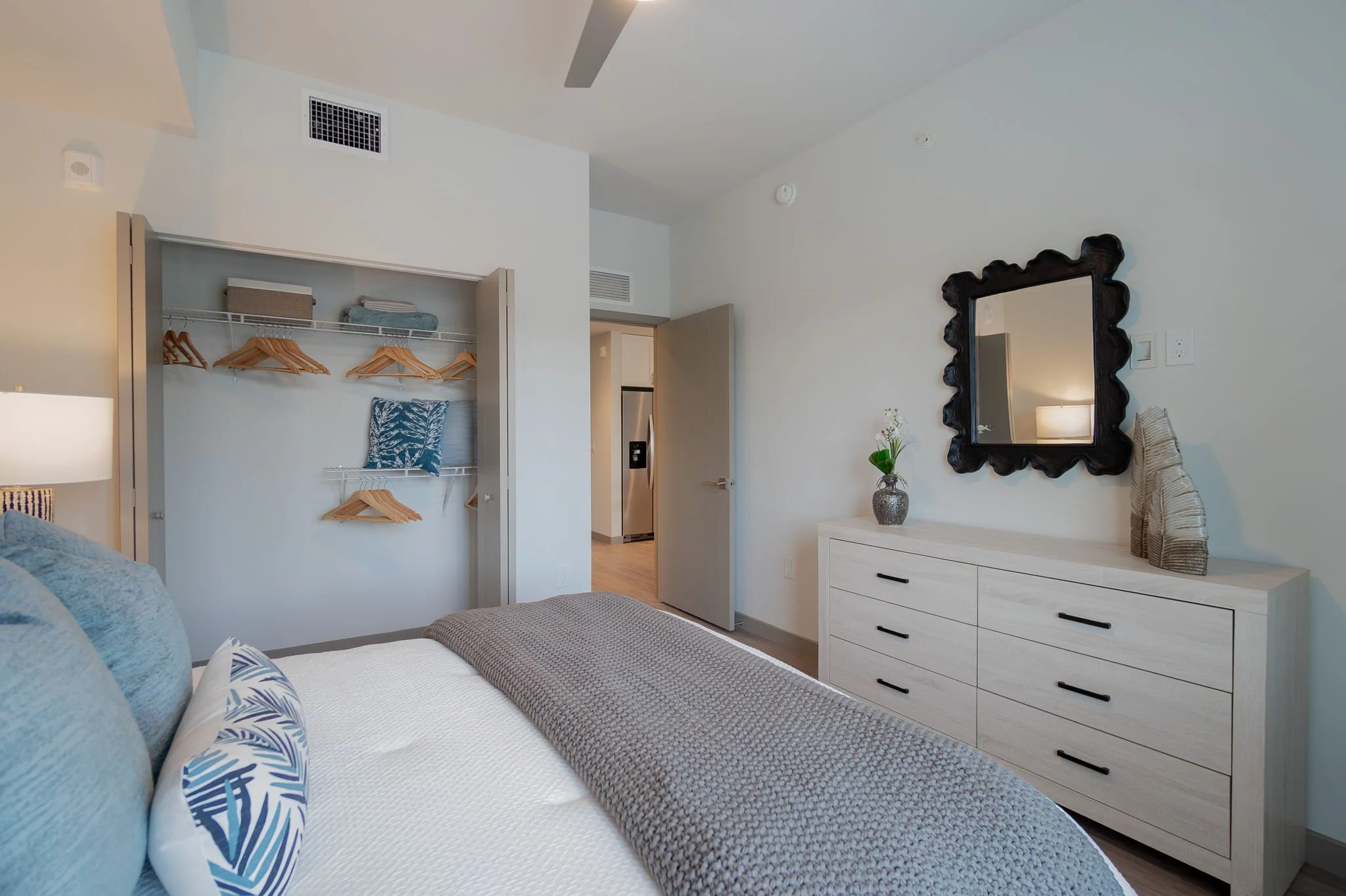 A bedroom with a bed, pillows, a bedside lamp, a dresser with a mirror, and an open closet with wooden hangers and some folded clothes.