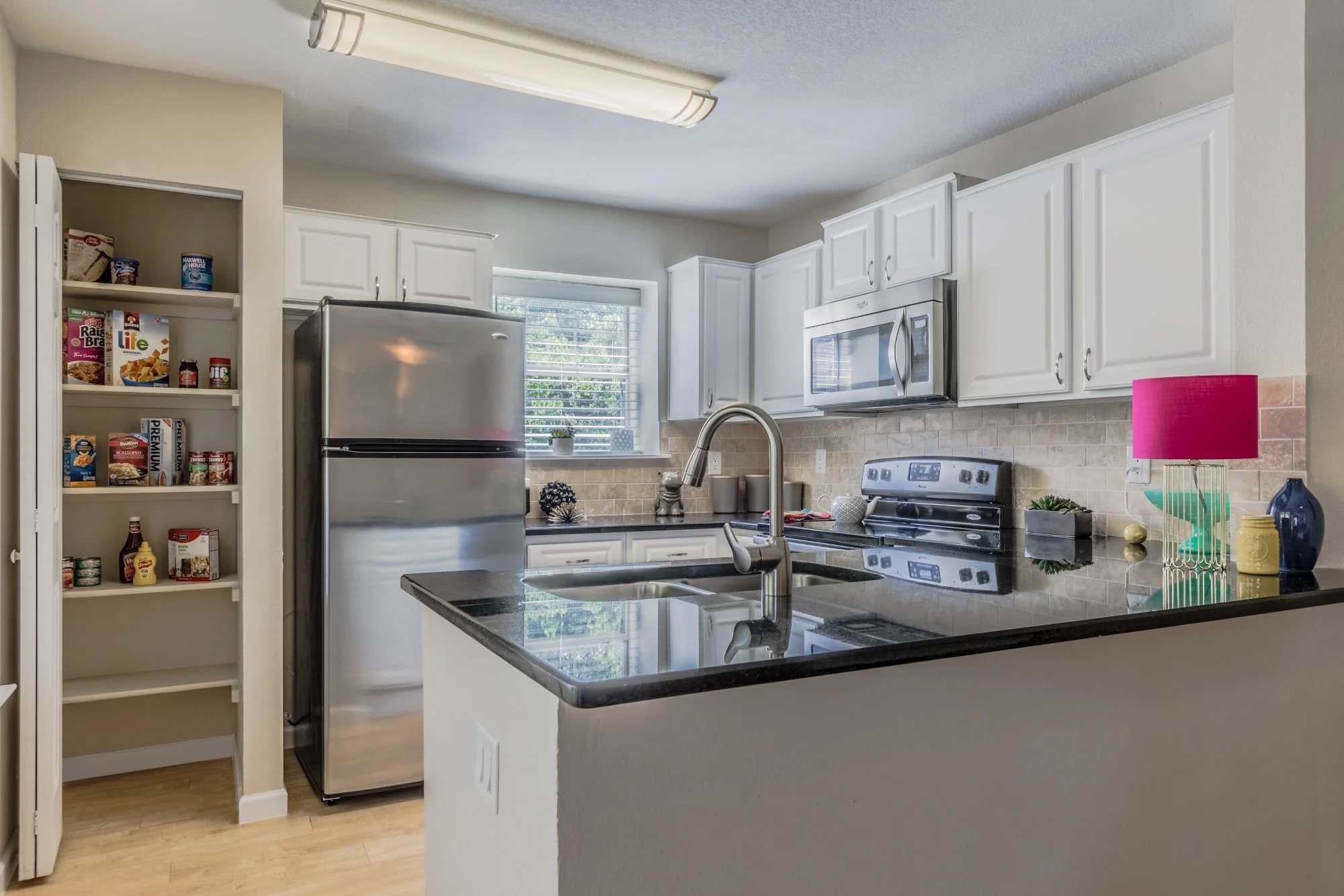 Kitchen with white cabinets, stainless steel refrigerator, microwave, and stove, black countertop island, beige tiled backsplash, and a window with blinds. Decor includes a pink lamp and various small plants and items.