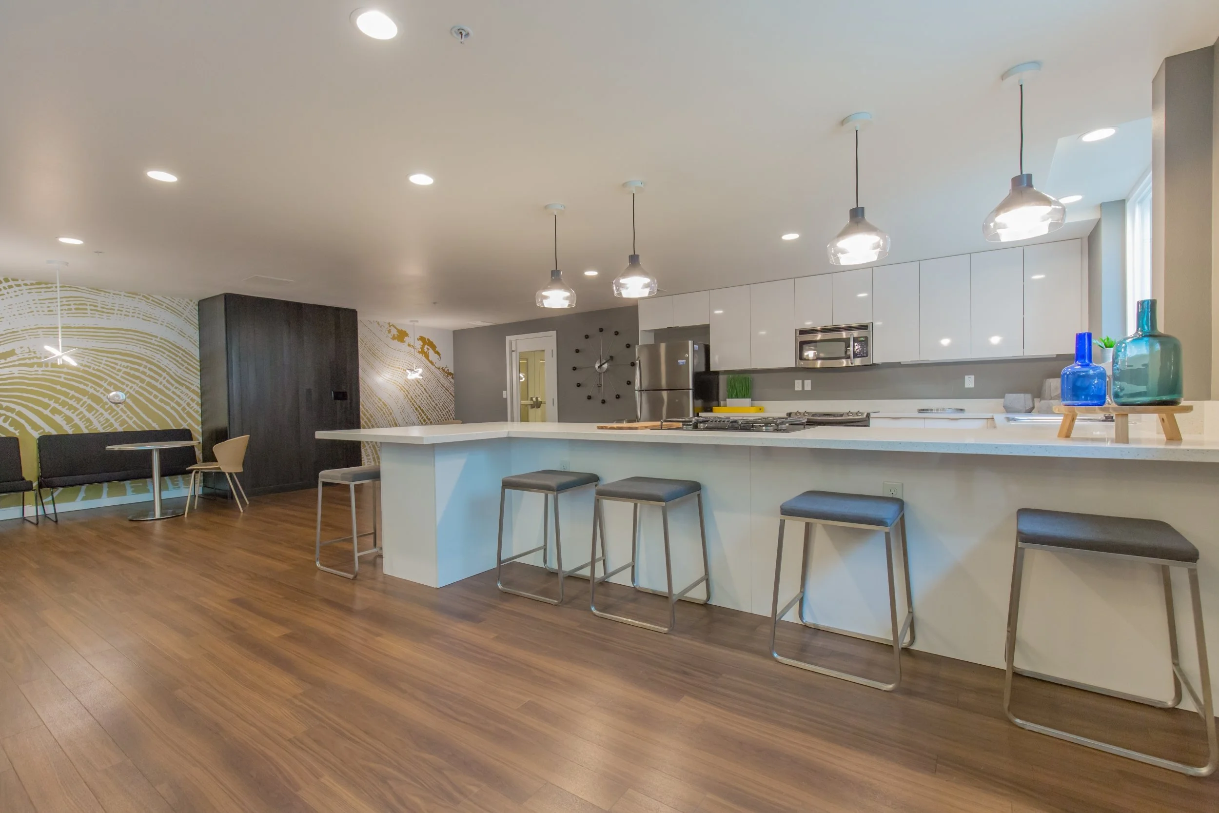 Modern open-concept kitchen with white cabinets, stainless steel appliances, a large island with seating, and pendant lights. Adjacent to a seating area with artwork on the wall and a dark wood accent wall.