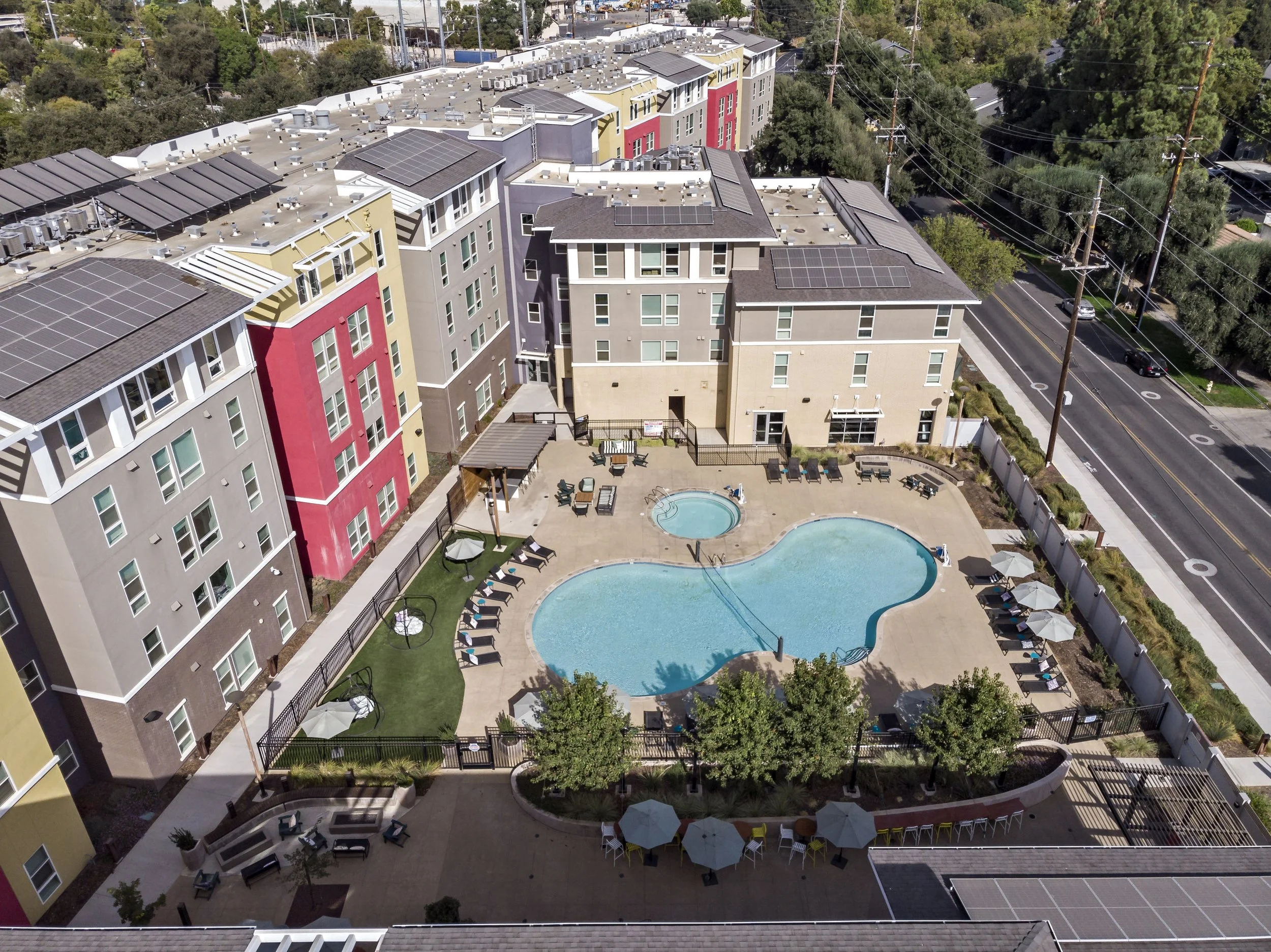 Aerial view of a modern apartment complex with a central swimming pool area, surrounded by multi-story residential buildings, outdoor seating, umbrellas, and greenery.