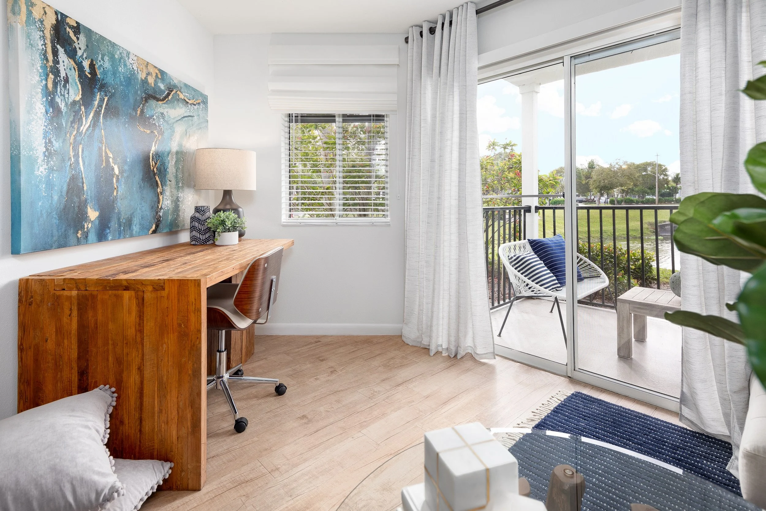 A living room with a wooden desk, a chair, a colorful abstract painting, a table lamp, potted plants, a sliding glass door with white curtains, a balcony with a chair and striped pillows, and a view of trees and a park outside.