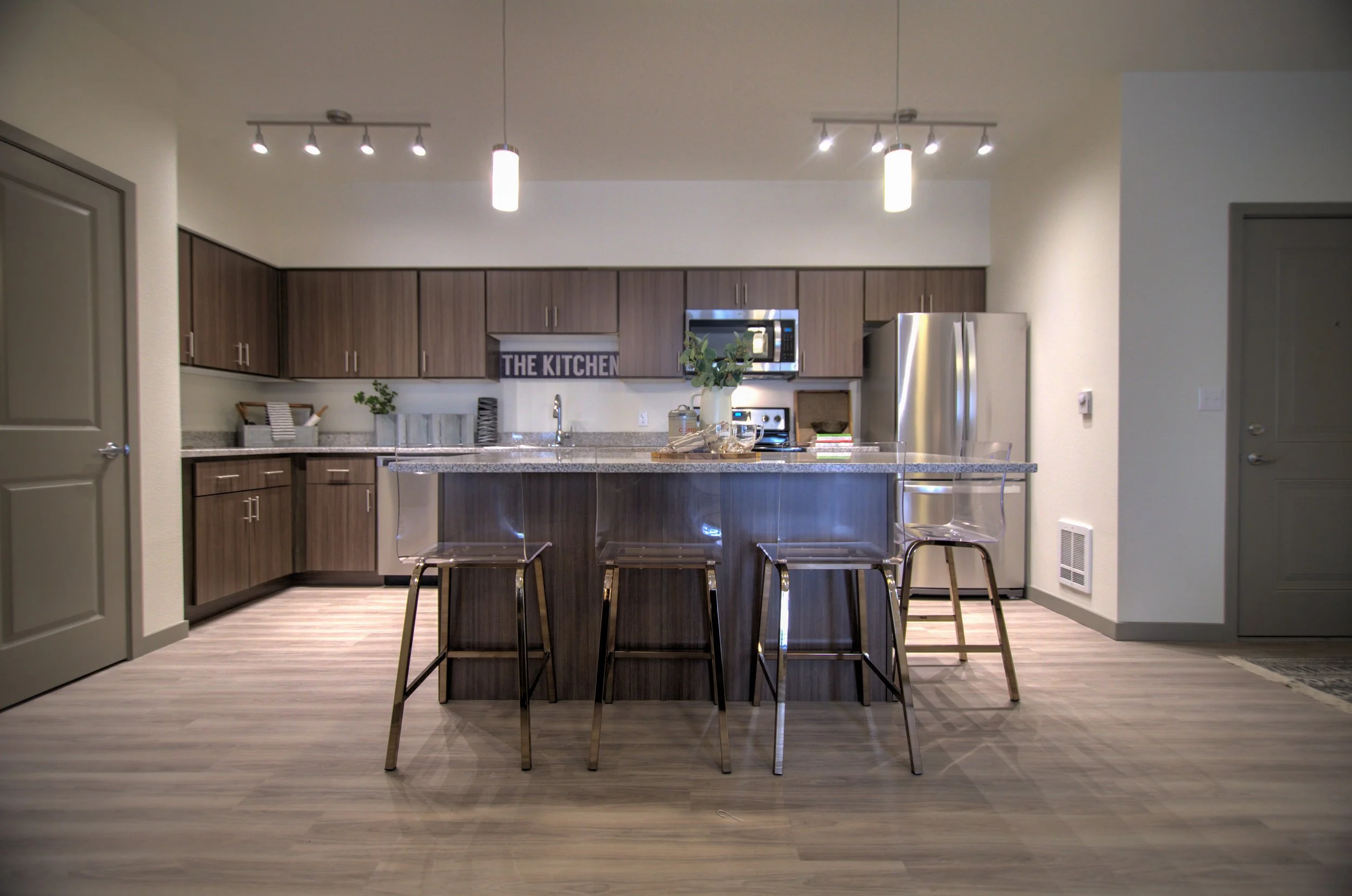 Modern kitchen with brown cabinetry, stainless steel appliances, a central island with four transparent chairs, and a sign that says 'THE KITCHEN' on the wall.