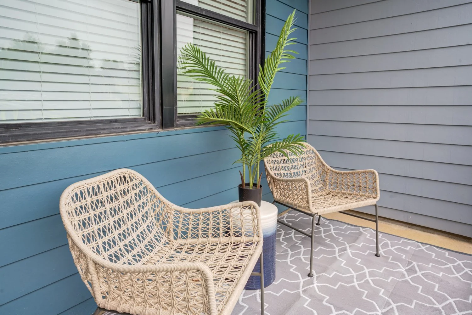 Outdoor porch area with two wicker chairs, a large potted plant, a patterned outdoor rug, and blue and gray siding on the house wall