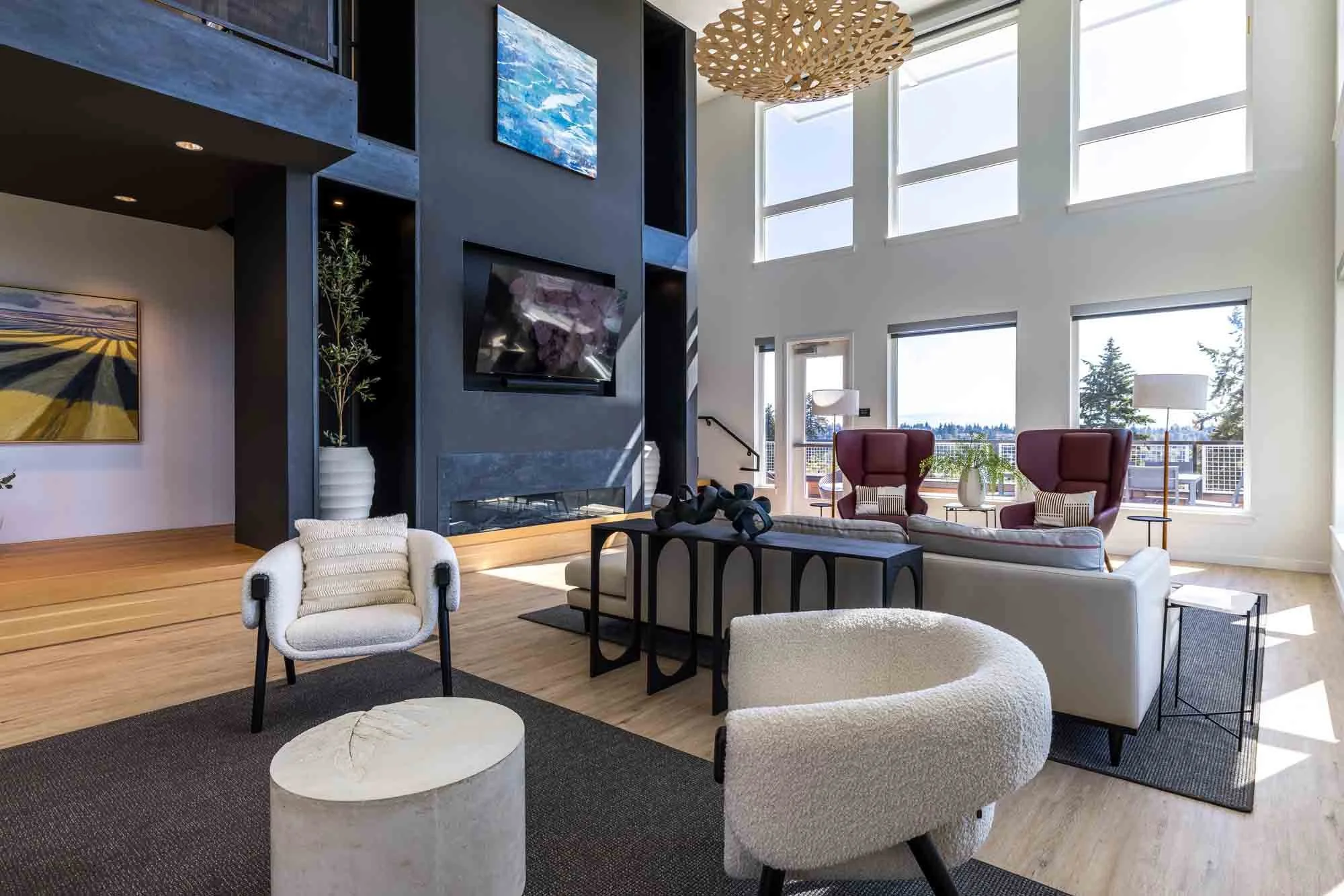 Modern living room with high ceilings and large windows, featuring a gray fireplace and wall-mounted TV, with contemporary furniture including white armchairs, a sofa, and red high-back chairs, decorated with lamps, plants, and artwork.