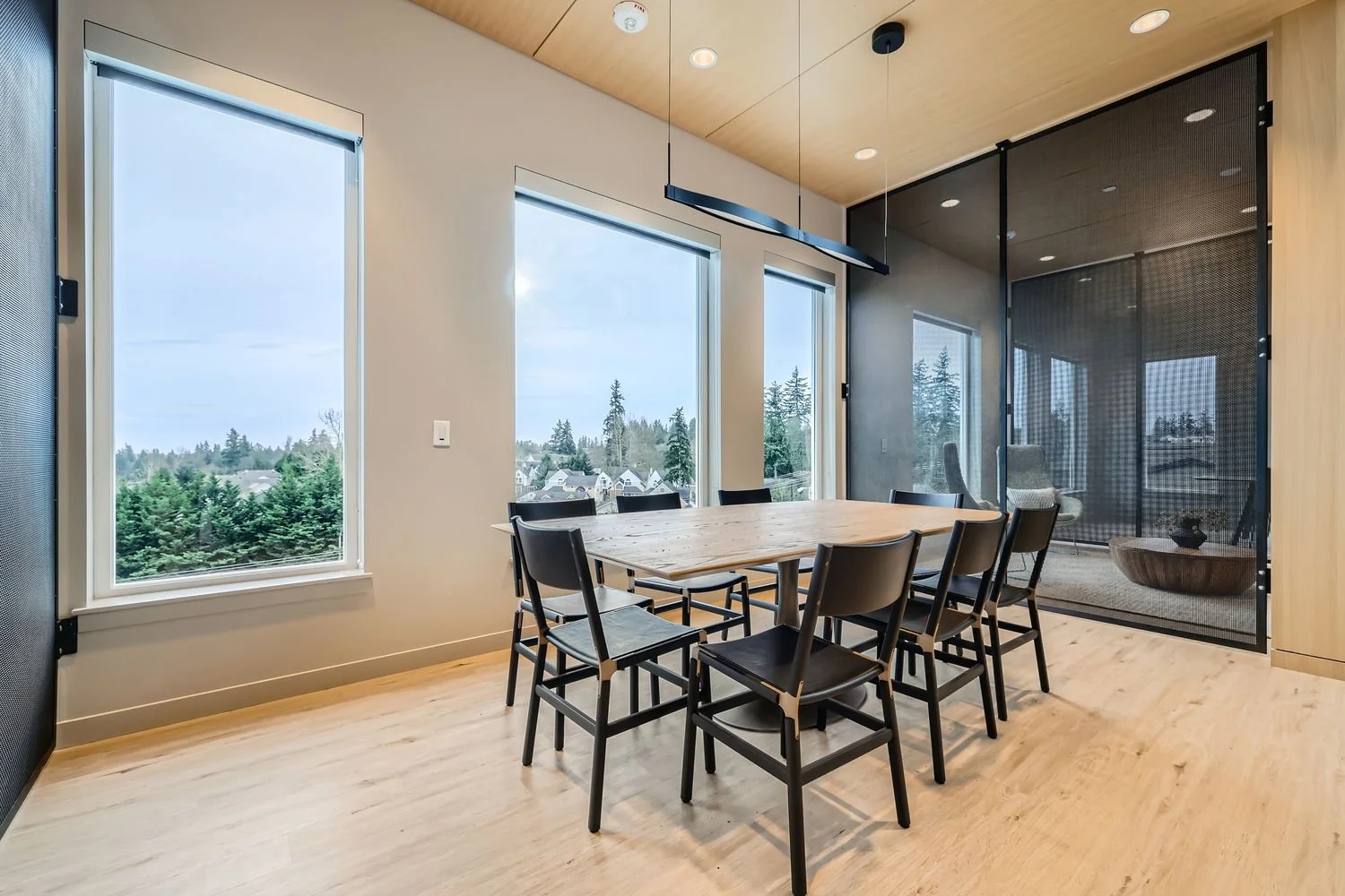 Modern dining room with wooden ceiling, large windows showing trees and neighborhood, wooden dining table, eight black chairs, and a glass wall with a mesh divider leading to sitting area.