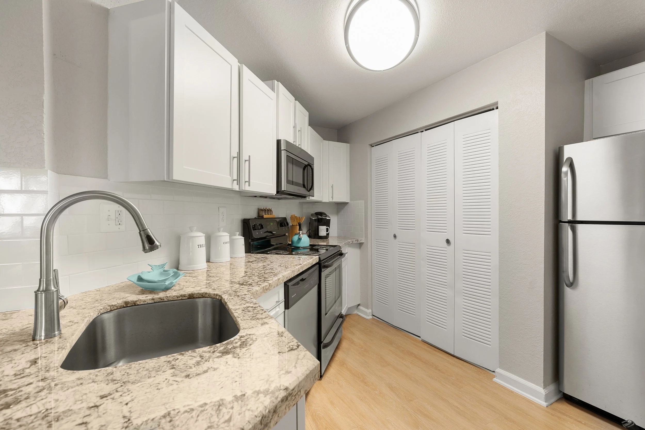 A modern kitchen with white cabinets, a granite countertop, stainless steel appliances, including a microwave, oven, dishwasher, and refrigerator. There is a small sink with a silver faucet, and on the counter are containers labeled 'Tea', 'Coffee', 