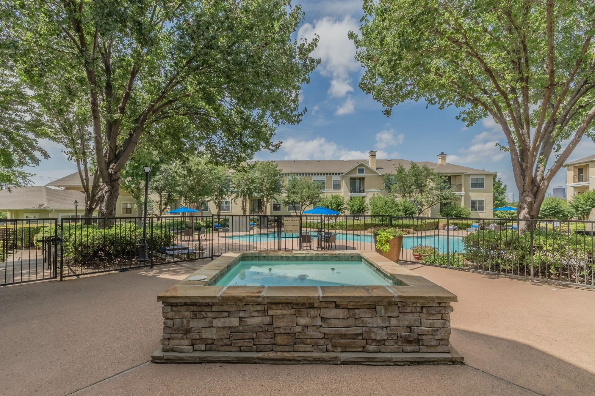 A swimming pool area with a hot tub in the foreground, surrounded by trees and apartment buildings in the background on a sunny day.