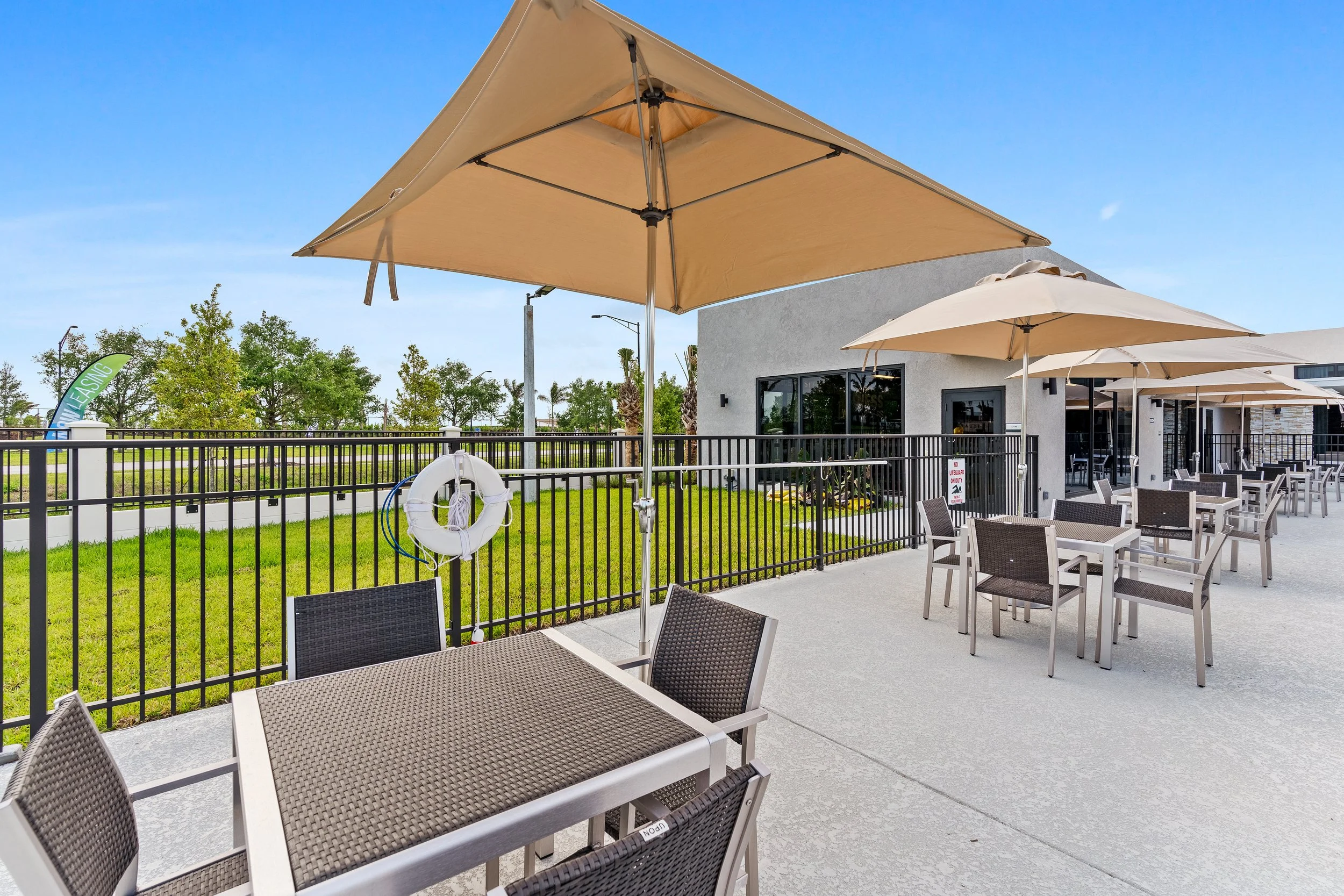 Outdoor patio with tables and chairs under large umbrellas, enclosed by a black metal fence, with a grassy area and trees in the background.