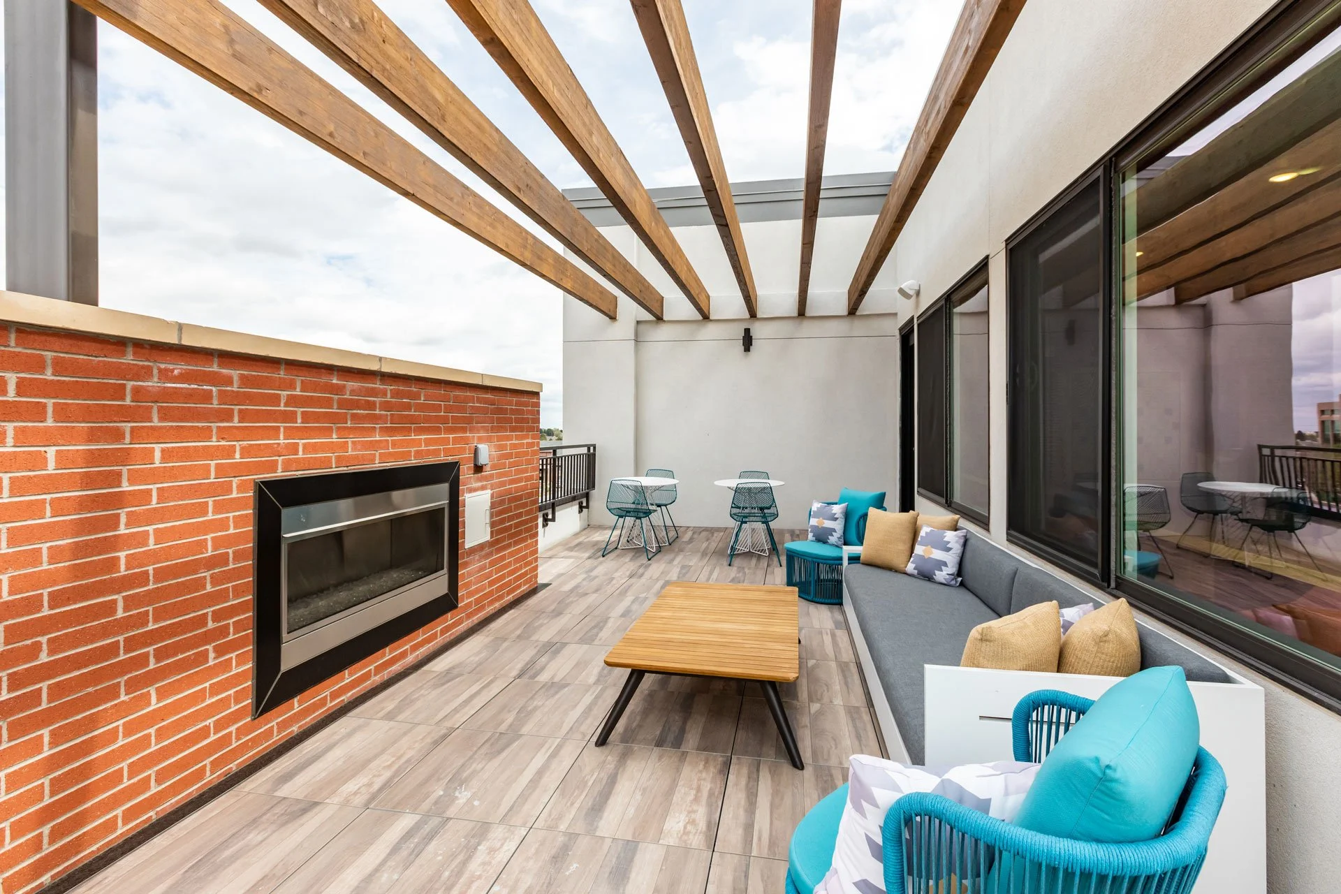 Outdoor rooftop patio with brick and white walls, wooden pergola, large windows, outdoor seating, blue and beige cushions, glass side table, and a gas fireplace.