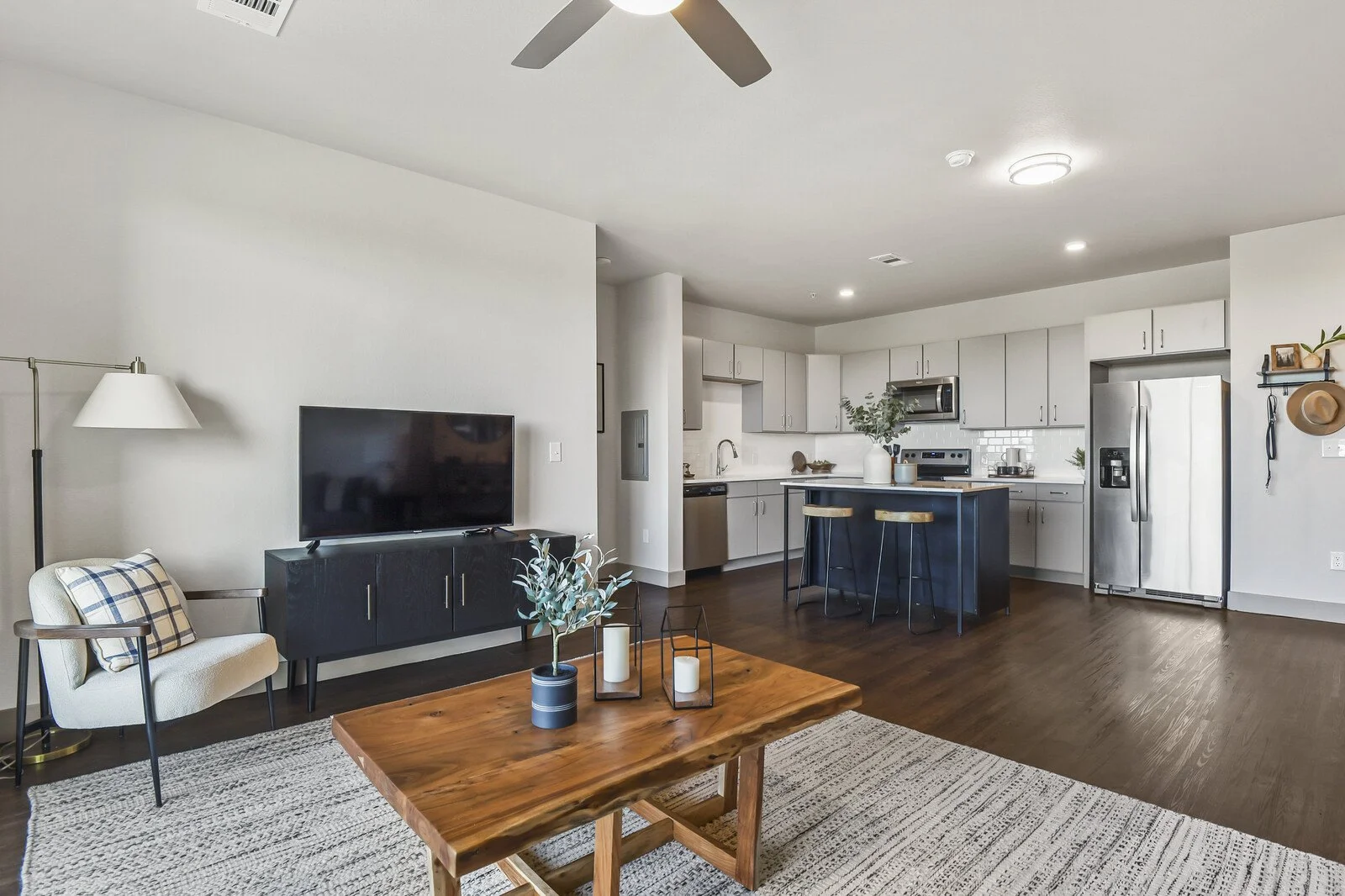 Living room with a TV, armchair, and wooden coffee table, open kitchen with white cabinets, stainless steel refrigerator, and small kitchen island with barstools.
