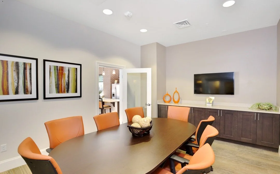 Modern conference room with a dark wood oval table, orange chairs, wall art, a wall-mounted TV, and decorative vases.