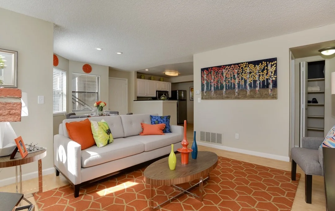 Living room with white sofa, colorful pillows, wooden coffee table with vases, orange patterned rug, and artwork on the wall, with a view into the kitchen.