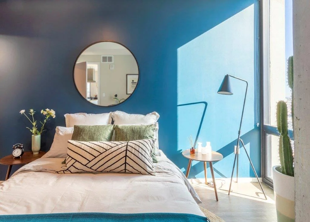 A modern bedroom features a blue accent wall, a round mirror, a bed with white and green pillows, a wooden side table with a tiny plant, glasses, aroma diffuser, and bottles, a blue standing lamp, and large windows with a cactus pot outside.