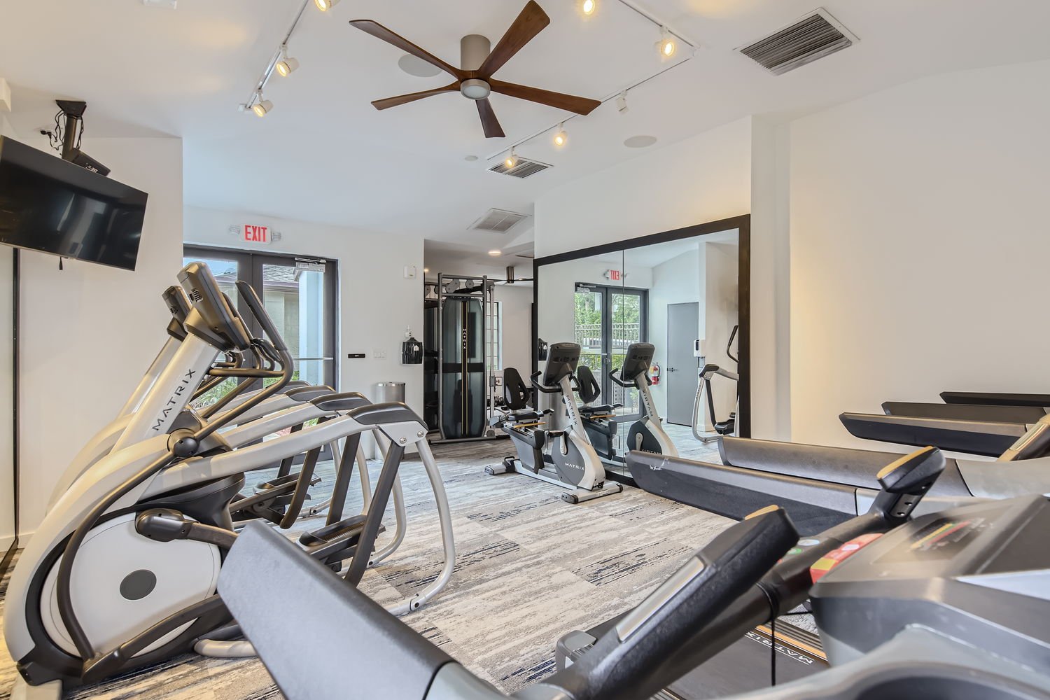 A modern gym with treadmills, stationary bikes, and elliptical machines, featuring a ceiling fan and track lighting.
