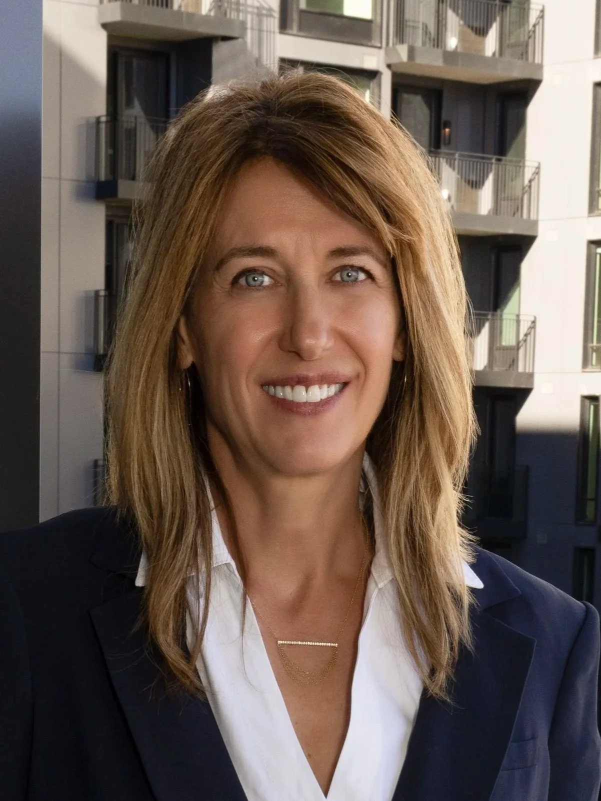 A woman with shoulder-length blonde hair, wearing a white collared shirt and a navy blazer, standing outside with modern apartment buildings in the background.