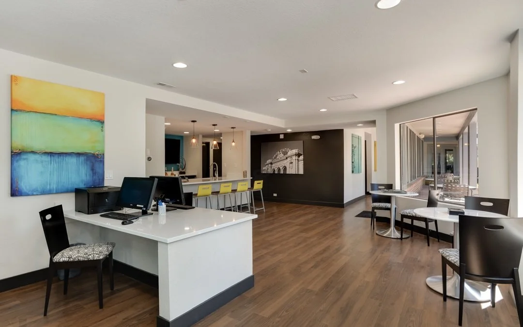 Modern coffee shop with white walls, wooden flooring, colorful abstract painting, black chairs, white tables, a computer station, and a bar counter with yellow barstools.