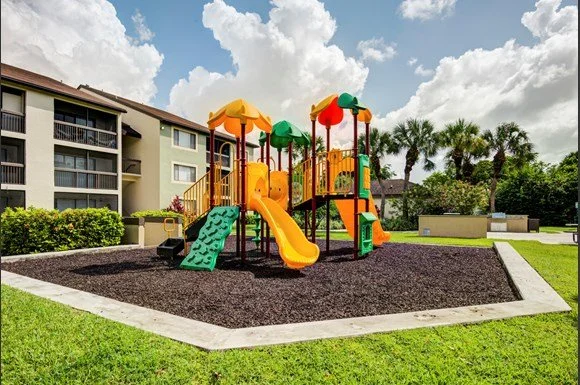Colorful playground with slides and climbing structures on black mulch, surrounded by green grass and apartment buildings, with trees and cloudy sky in the background.