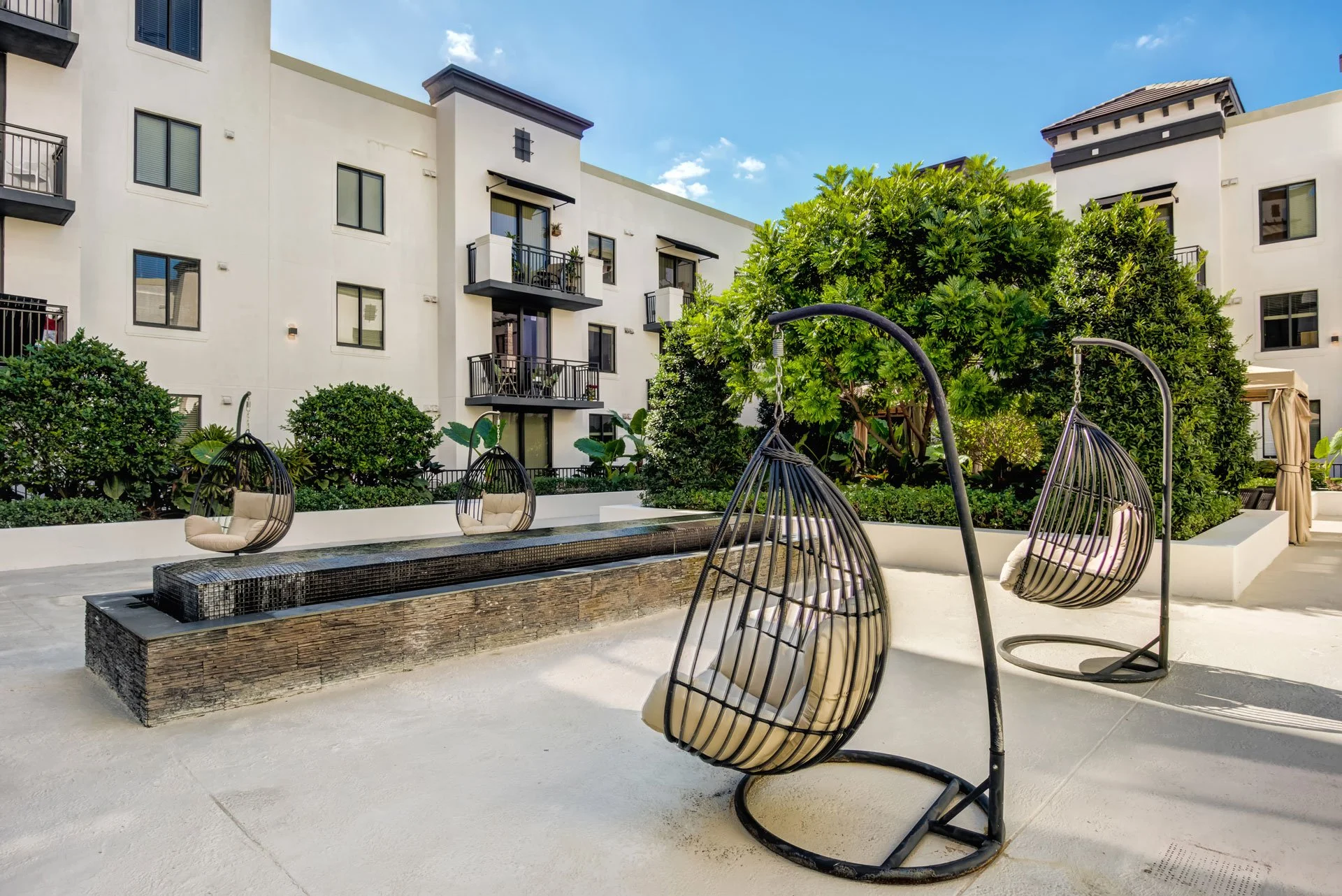 Outdoor patio with two modern hanging egg chairs with beige cushions, a rectangular water feature, lush green bushes and trees, and apartment buildings in the background under a blue sky.